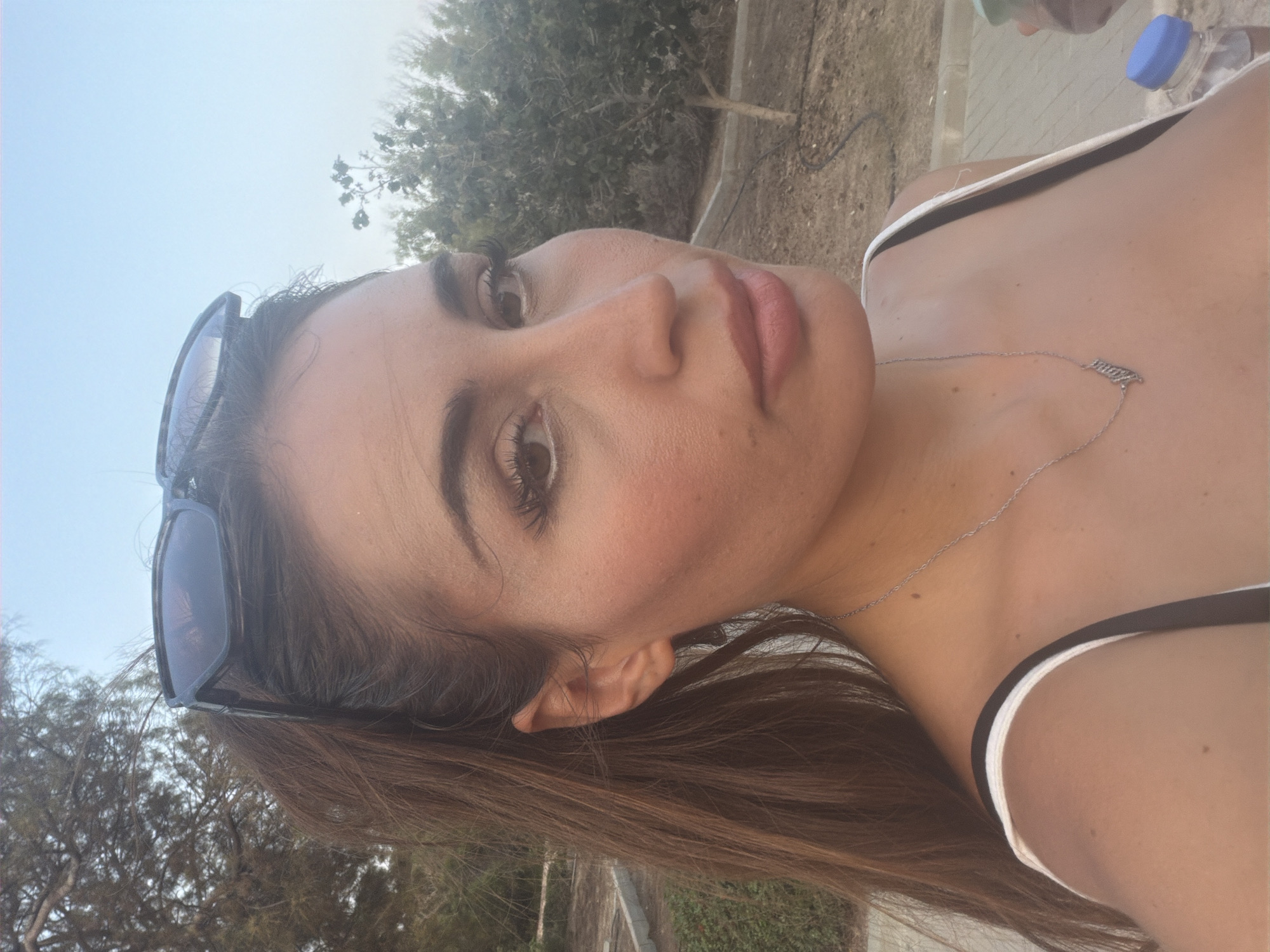 This media file features a close-up, rotated selfie of a young woman. She is outdoors, with trees and a paved area visible in the background, suggesting a park or a scenic outdoor location. The sky is bright and clear, indicating it is daytime with fair weather. She is wearing sunglasses on her head and a white tank top with black straps. A delicate silver necklace with a small, intricate pendant rests on her collarbone. Her hair is long and brown, framing her face. Her expression is relaxed and pleasant. In the background, a plastic bottle with a blue cap is visible, along with another person's arm and shoulder, also suggesting an outdoor gathering. The scene appears to be set in Pervolia, Cyprus. There is no visible text in the image.