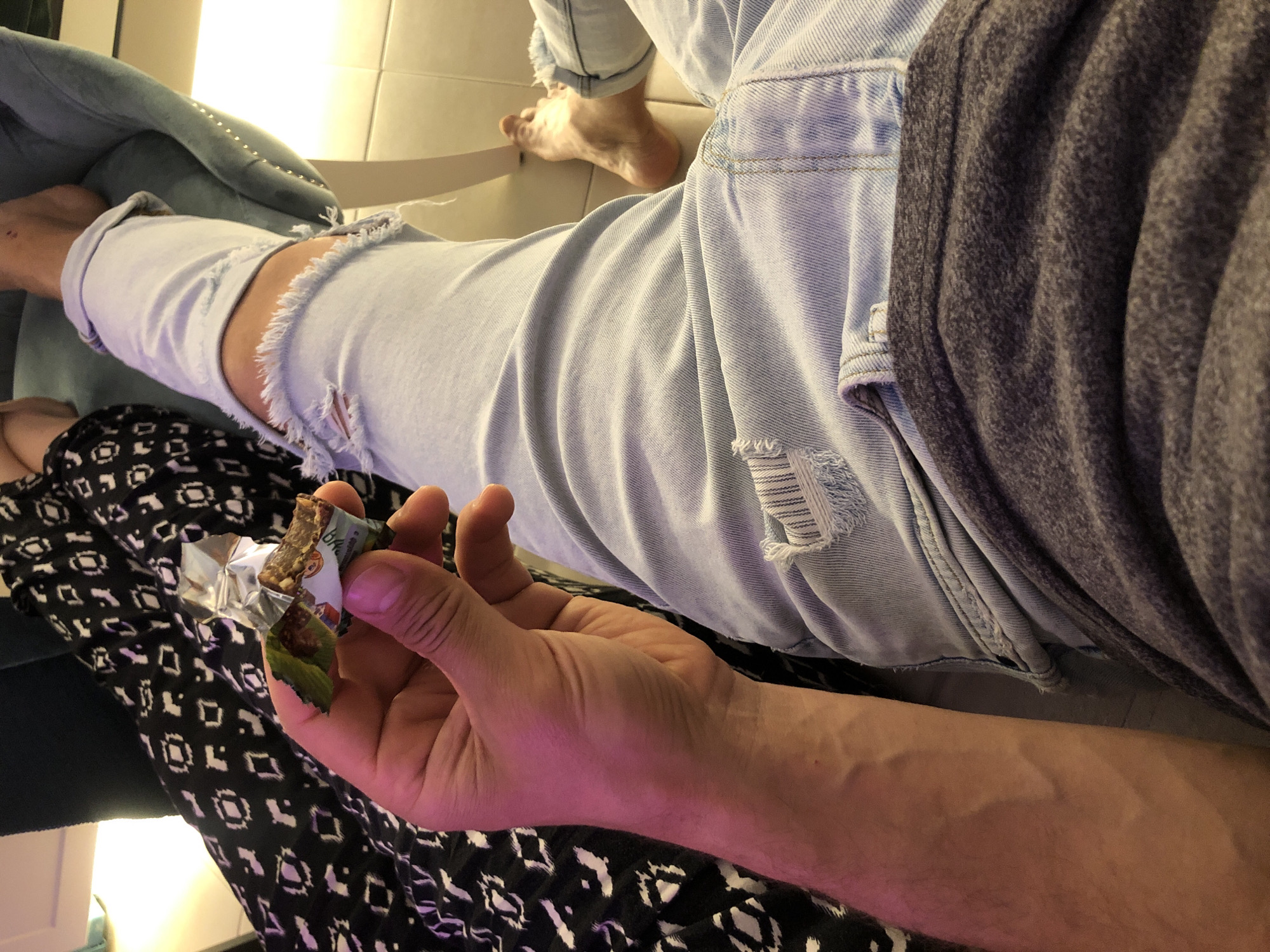 An interior scene in Saint Petersburg, Russia, captures a close-up, downward perspective of two individuals relaxing. In the foreground, a person's bare right forearm and hand, exhibiting prominent veins, holds a partially unwrapped snack bar. The snack bar's packaging is silver, green, and white, with the Russian word "Ярко" (Yarko) visible, indicating a Russian product and thus reinforcing the Saint Petersburg location. This individual is wearing light blue distressed denim jeans with visible rips at the knee and upper thigh, along with a dark grey t-shirt or sweater. Their bare right foot is partially visible, resting on a light-colored tiled floor. To the left, a second person's legs are visible, clad in black fabric with a white geometric pattern. One of their bare feet is positioned on a light blue upholstered chair, which features a row of metallic studs along its edge. The setting is indoors, illuminated by warm artificial light.