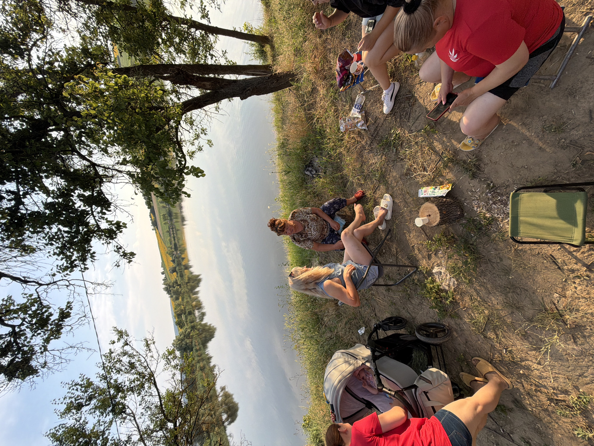 The image depicts a group of people, predominantly women and a baby, gathered by a body of water, likely a lake, on a sunny day. The scene appears to be a picnic or a casual outdoor gathering.
**People:**
* One woman in a red t-shirt and denim shorts is seen from the back, pushing a stroller.
* A baby is visible in the stroller.
* A woman with blonde hair, wearing a denim dress, is sitting in a folding chair.
* Another woman, with dark hair pulled up, is wearing a leopard-print top and patterned pants. She is seated on the ground.
* Two other women are partially visible, one in a red shirt, sitting in chairs, and another with blonde hair, also in a chair, looking at her phone.
**Objects:**
* A baby stroller is present.
* Several folding chairs are set up.
* Bags of snacks and drinks, including a bottle of water and a red cup, are scattered on the ground.
* A small wooden log is used as a surface for a cup and a juice box.
* A mobile phone is being held by one of the women.
* A green folding chair is visible in the foreground.
**Environment:**
* The location is beside a calm body of water with reflections of the sky and trees on its surface.
* The terrain is uneven and covered with grass and dry soil.
* Trees with green foliage line the edge of the water, providing shade.
* In the background, a rolling landscape with fields of sunflowers and other vegetation is visible under a clear blue sky with some clouds.
**Actions and Interactions:**
* The group appears to be relaxed and enjoying their time outdoors.
* One woman is tending to the stroller.
* Some individuals are seated, while others are interacting or looking at their phones.
**Location:**
The scene is situated near a lake, with agricultural fields in the background, consistent with the rural landscape of Pănășești, Moldova. The presence of sunflowers in the background is a strong indicator of this region.