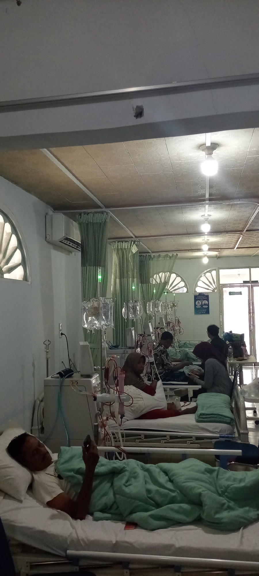 The image shows a group of patients undergoing dialysis treatment in a hospital ward. Multiple dialysis machines and IV fluid bags are visible, along with patients lying on beds, some covered with blankets and others hooked up to medical equipment. The room appears to be a ward with multiple beds arranged side-by-side, separated by green curtains. A window with an arched design and an air conditioning unit are present on the wall. A sign with the word "KELUAR" (exit in Indonesian) is visible near a doorway, suggesting the location is a public facility. The overall impression is one of a clinical setting dedicated to patient care, likely a dialysis unit within a hospital in Lhokseumawe, Indonesia. The lighting appears to be from overhead fluorescent fixtures.