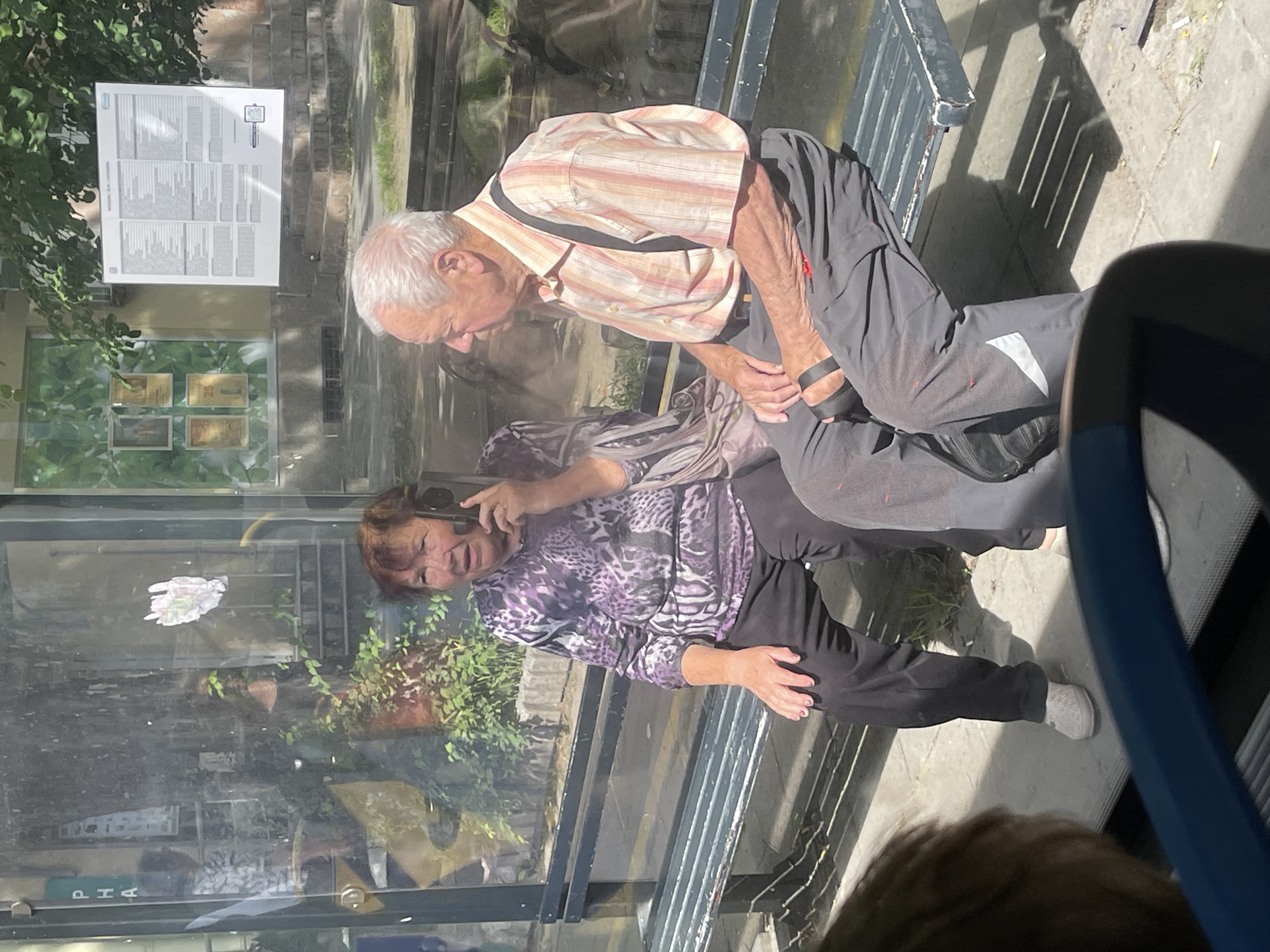 This full-frame image, captured from within a vehicle, depicts an outdoor scene in the city of Unknown, Unknown, likely at a bus stop or public waiting area on a sunny day. Two elderly individuals are seated on a dark grey, slatted metal bench.
On the left, an elderly woman with short, reddish-brown hair, wearing a purple and black animal-print long-sleeved top and black trousers, is holding a black smartphone to her right ear, seemingly engaged in a conversation with her mouth slightly open. Her fingernails are painted pink, and she wears light-colored slip-on shoes.
To her right, an elderly man with short, white hair is seated. He wears a short-sleeved, collared shirt with vertical stripes in shades of beige, orange, and white, dark grey trousers, and black suspenders. He has dark sandals on his feet, and a small black bag is attached to his right leg. He is looking to his left, away from the woman.
The background features a building with large glass windows reflecting trees and several framed posters, one partially legible with "PHA" vertically printed. A large white sign with extensive black text and a QR code is mounted above, partially obscured by foliage. The ground is concrete, showing distinct shadows from the bench slats, indicating strong sunlight. Several cigarette butts are visible on the pavement near the man's feet. A blurred blue object, consistent with a vehicle's interior, occupies the bottom foreground of the image.