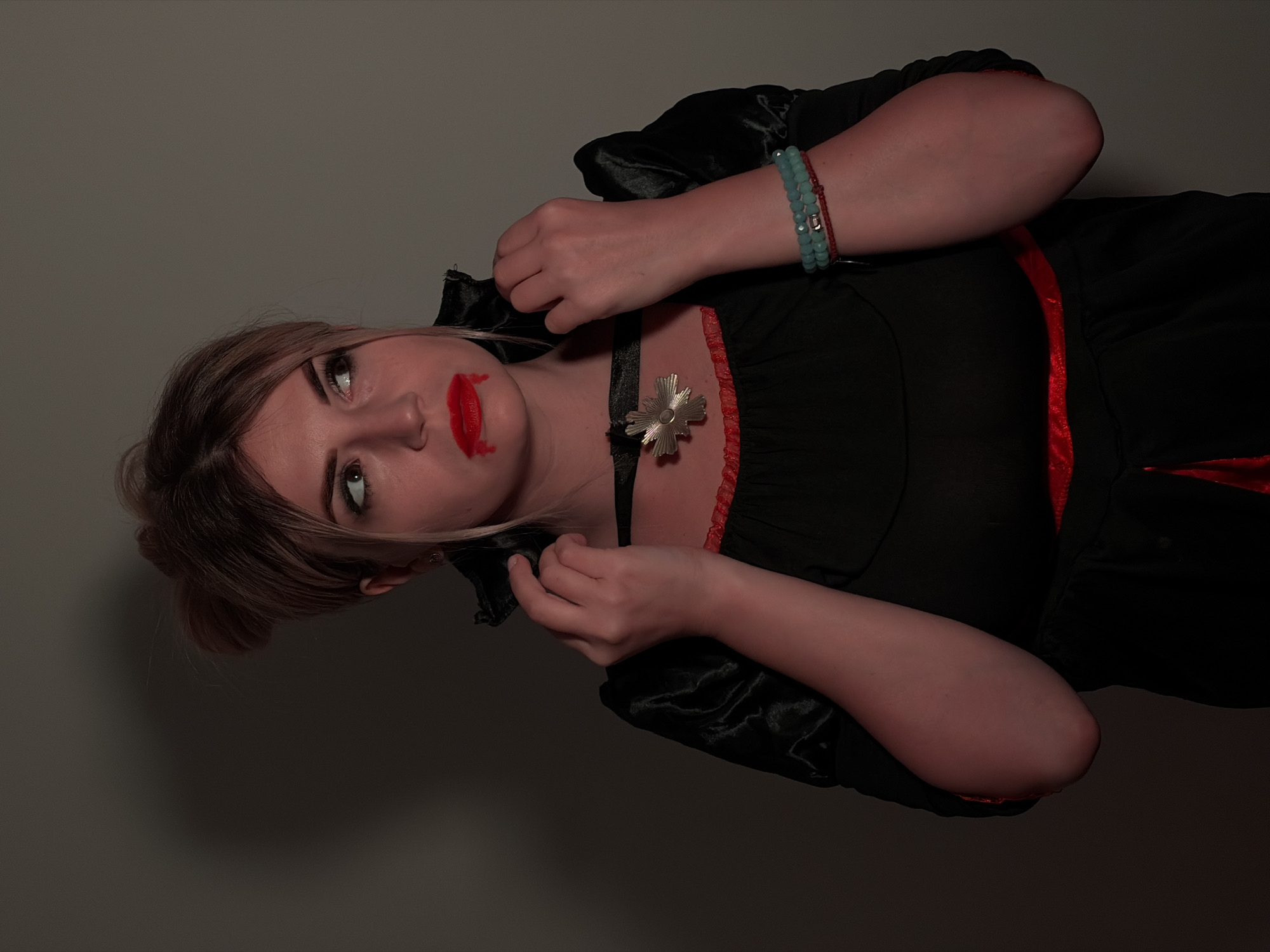 A young woman dressed in a vampire costume is depicted in a portrait-style photograph, rotated 90 degrees clockwise. She is positioned against a plain, dark grey wall, suggesting an indoor setting.
The woman has light brown hair with lighter highlights. Her makeup includes prominent eyeliner and bright red lipstick, which is intentionally smudged below her lower lip to simulate dripping blood, enhancing the vampiric theme. She wears a black dress with red trim along the neckline and possibly the sleeves, featuring puffed shoulders. A black choker with a silver, cross-shaped pendant is visible around her neck. On her left wrist, she wears a turquoise beaded bracelet and what appears to be a red string bracelet.
Her gaze is directed upwards and slightly to the left, conveying a dramatic or intense expression. Her hands are raised towards her neck and collar, as if adjusting her costume or posing. The lighting is somewhat dim, casting subtle shadows and contributing to a mysterious atmosphere.
The image captures a theatrical pose, indicative of a costume event like Halloween or a themed party. There are no visible texts, and no specific location cues are present within the frame itself. The overall mood is one of playful horror, consistent with a vampire portrayal.