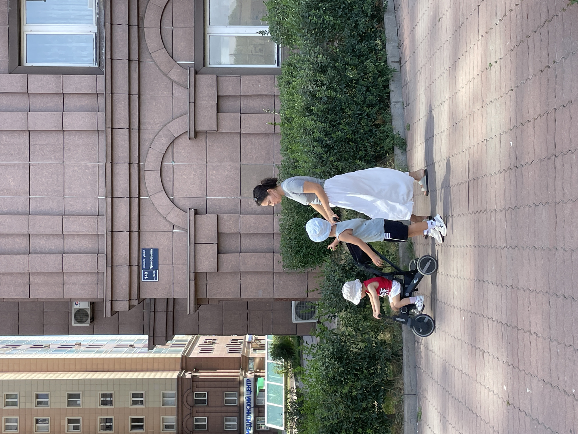 The image shows a woman and two children on a sidewalk in front of a modern building. The woman is wearing a gray t-shirt and a long white skirt, and is pushing a stroller. A young boy in a light blue t-shirt and black shorts is standing next to her, looking at the stroller. A smaller child wearing a red shirt and white shorts is sitting in the stroller, which is a tricycle-stroller hybrid. The building behind them is made of pinkish-brown tiles, with arched window frames and a street sign that reads "140 улица Прокофьева" in Russian, indicating the location is Prokofieva Street in Almaty, Kazakhstan. To the left of the building, a lighter colored, taller building with many windows is visible, and further left, a sign reads "МЕДИЦИНСКИЙ ЦЕНТР" (Medical Center). Lush green bushes line the sidewalk in front of the buildings, and the sidewalk itself is paved with reddish-brown interlocking bricks. The scene appears to be a bright, sunny day.