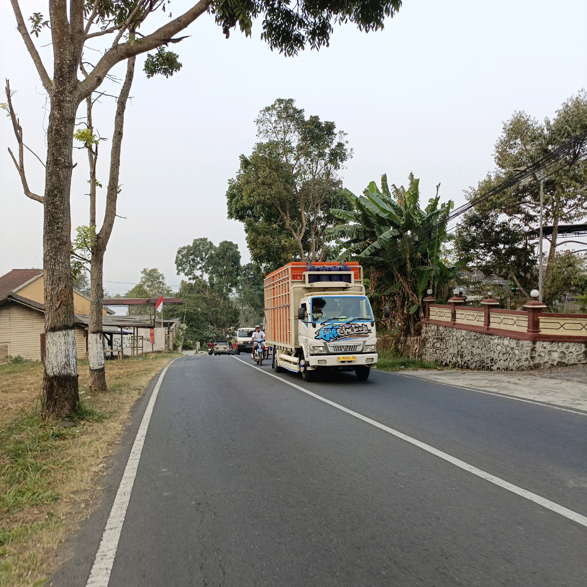 A white truck with an orange cage on its back is driving on a paved road in Sumogawe, Indonesia. The truck has blue accents and graphics on the front. A person is riding a motorcycle in front of the truck, and a car is behind it. The road is lined with trees on the left side and a stone wall with decorative elements on the right. The sky is overcast.