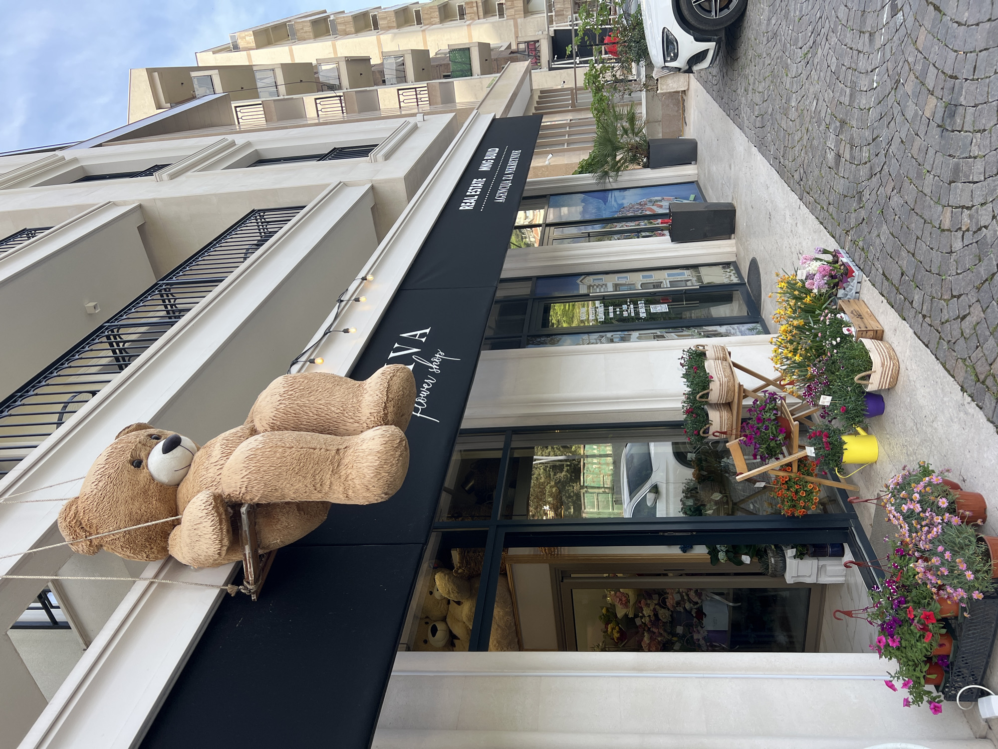 The image depicts the facade of a flower shop named "Rena Flower Shop" in Budva, Montenegro. The building is a modern construction with a white facade. A large teddy bear is suspended from the black awning of the shop. The awning also bears the sign "Real Estate - MMG Build" and "Agencija za Nekretnine". The shopfront features large glass windows, displaying the floral arrangements within. Outside the shop, various potted plants and flower arrangements are displayed on wooden stands and the ground. The street is paved with cobblestones, and a white car is partially visible. In the background, a taller building with multiple balconies is visible under a blue sky.