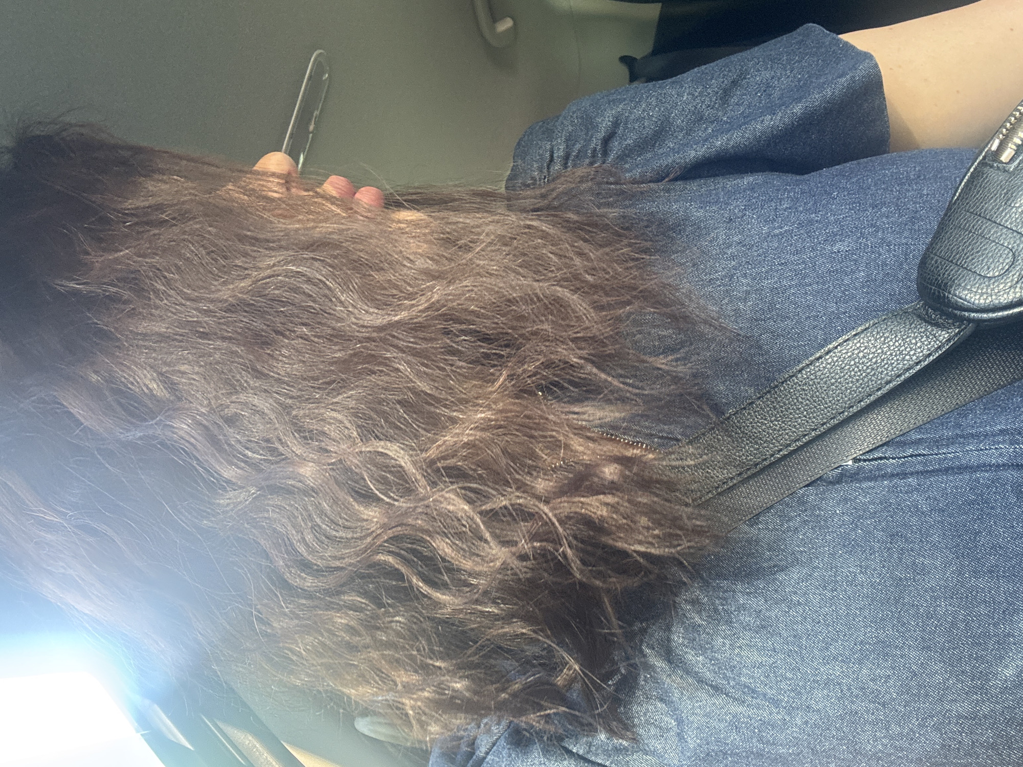 A person with long, wavy, dark brown hair is viewed from behind and to the side, inside a vehicle. The person is wearing a denim shirt with puffed sleeves. A black crossbody bag with textured leather and a zipper is visible across the person's chest. The interior of the vehicle appears to be the passenger compartment, with a glimpse of the headliner and a grab handle. Sunlight streams in from the left side of the frame, causing lens flare and washing out parts of the image. The background is indistinct and dark. There is no indication of the specific location within the city of Dnipro, Ukraine.