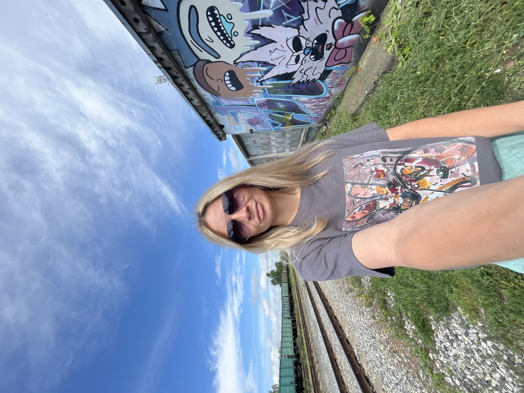 A blonde woman wearing sunglasses and a t-shirt with a colorful abstract design takes a selfie. She is standing next to a graffiti-covered wall, with train tracks and a train in the background. The sky is blue with scattered white clouds. This scene appears to be an industrial area, likely near a railway yard. The graffiti style suggests a vibrant, urban art presence.