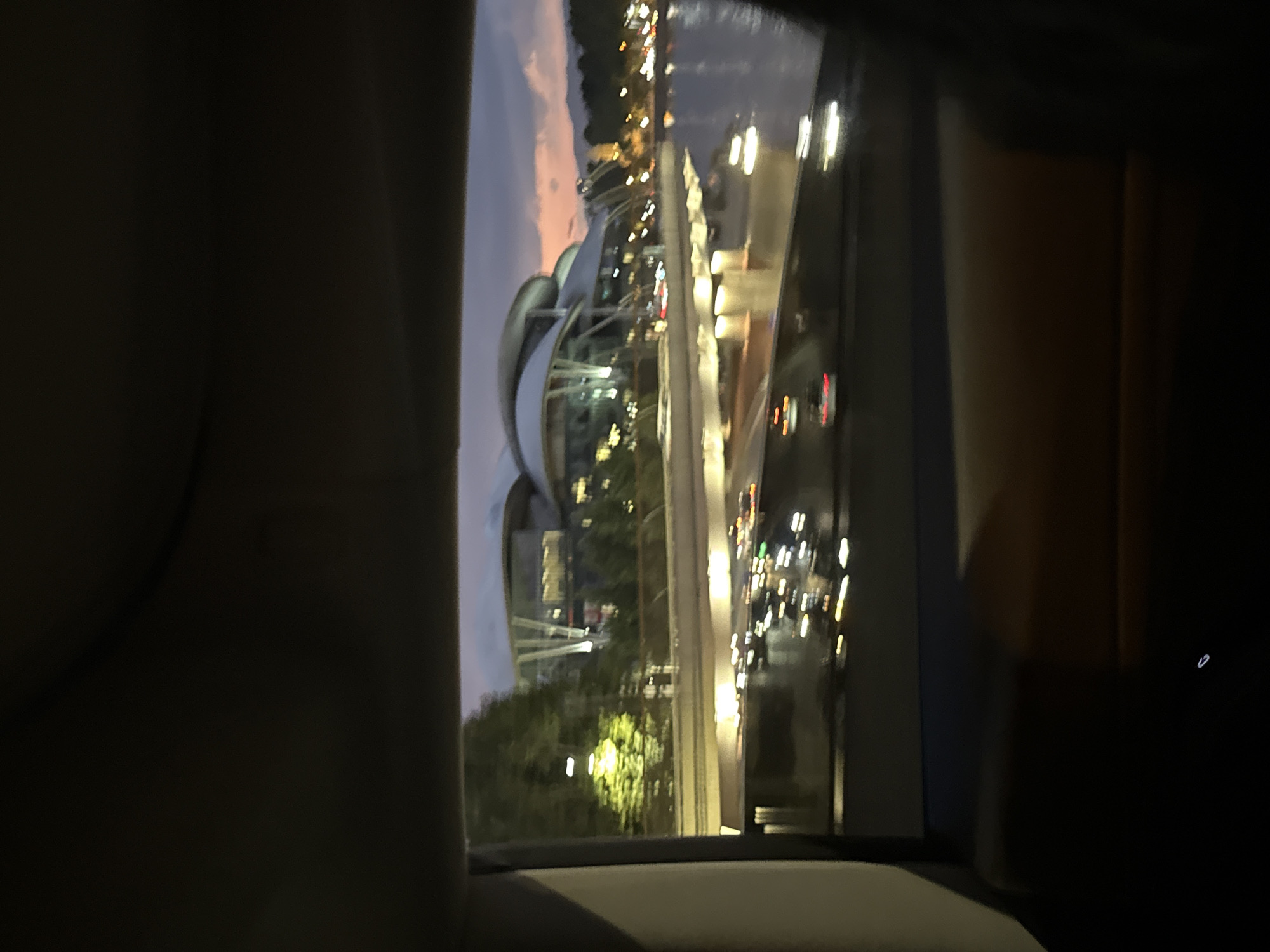 The image shows a view from inside a vehicle, likely at dusk or night, looking out at a cityscape. The scene is dominated by a modern architectural structure with a curved, metallic roof, illuminated by numerous lights. Adjacent to this building, there is a body of water, possibly a river, reflecting the lights of the city.
On the right side of the frame, there are blurred streaks of light, indicating movement, suggesting the vehicle is in motion. These lights also illuminate the interior of the car, revealing the brown upholstery of a seat and a dark dashboard. The sky exhibits a gradient of colors, transitioning from a dusky pink and orange near the horizon to a deep blue or purple higher up, typical of twilight.
The overall impression is that of a vibrant urban environment at night, seen from the perspective of a passenger. The lights create a festive and dynamic atmosphere, while the reflections on the water add depth and visual interest. The blurriness of the image suggests it was taken with a moving camera, possibly from a car window. There is no visible text in the image. The location context suggests this is a scene from Tbilisi, Georgia.