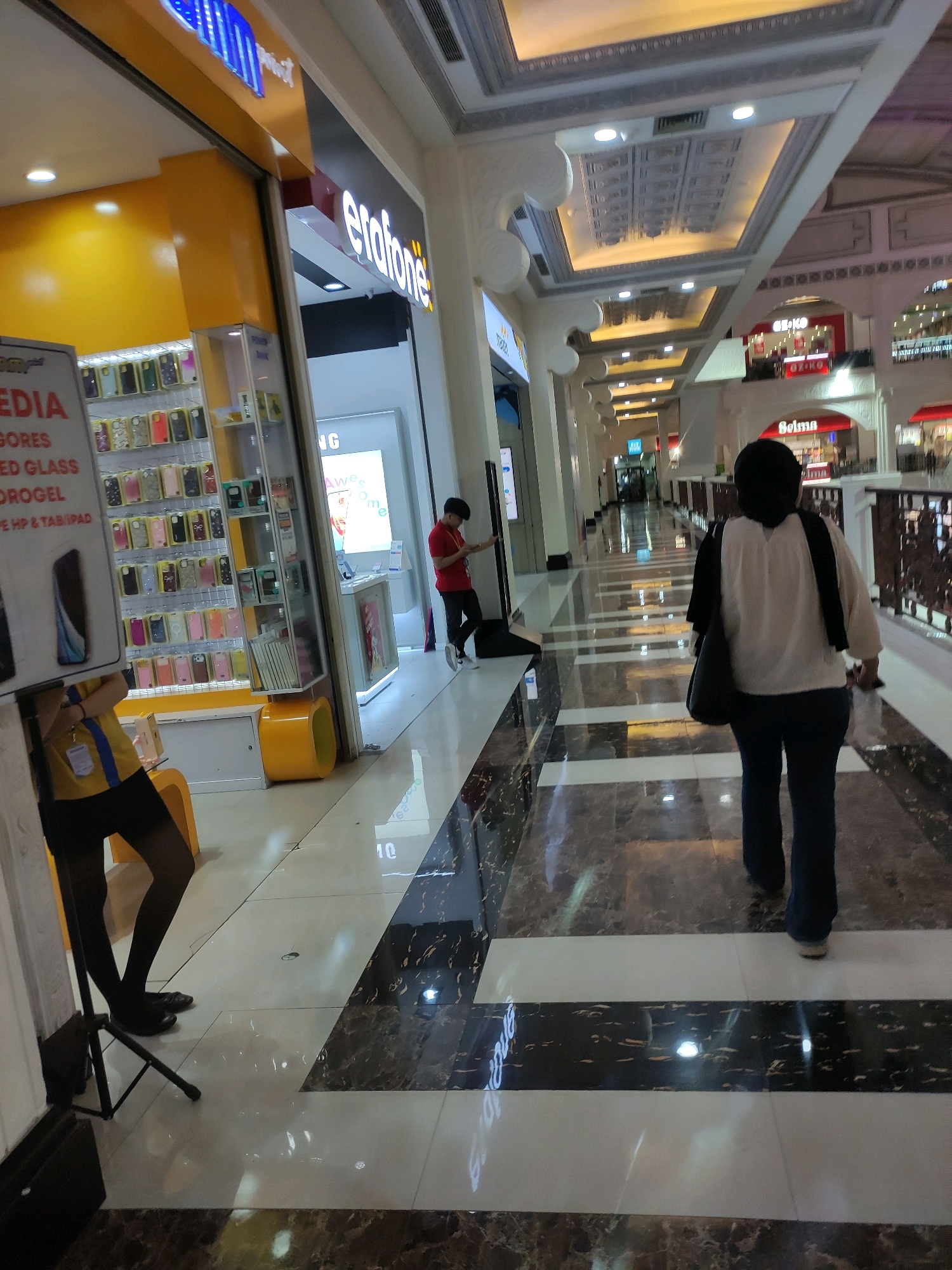 The image depicts an interior view of a shopping mall in Sinduadi, Indonesia. In the foreground, a person walks away from the camera, wearing a white long-sleeved shirt, dark pants, and a black headscarf. To the left, an Erafone store is visible, displaying various phone accessories. Another person in a red shirt stands inside the store, looking at a phone. The mall's interior features marble flooring with a pattern of black and white stripes, decorative ceilings, and well-lit aisles. The overall scene captures a typical day in a commercial establishment.