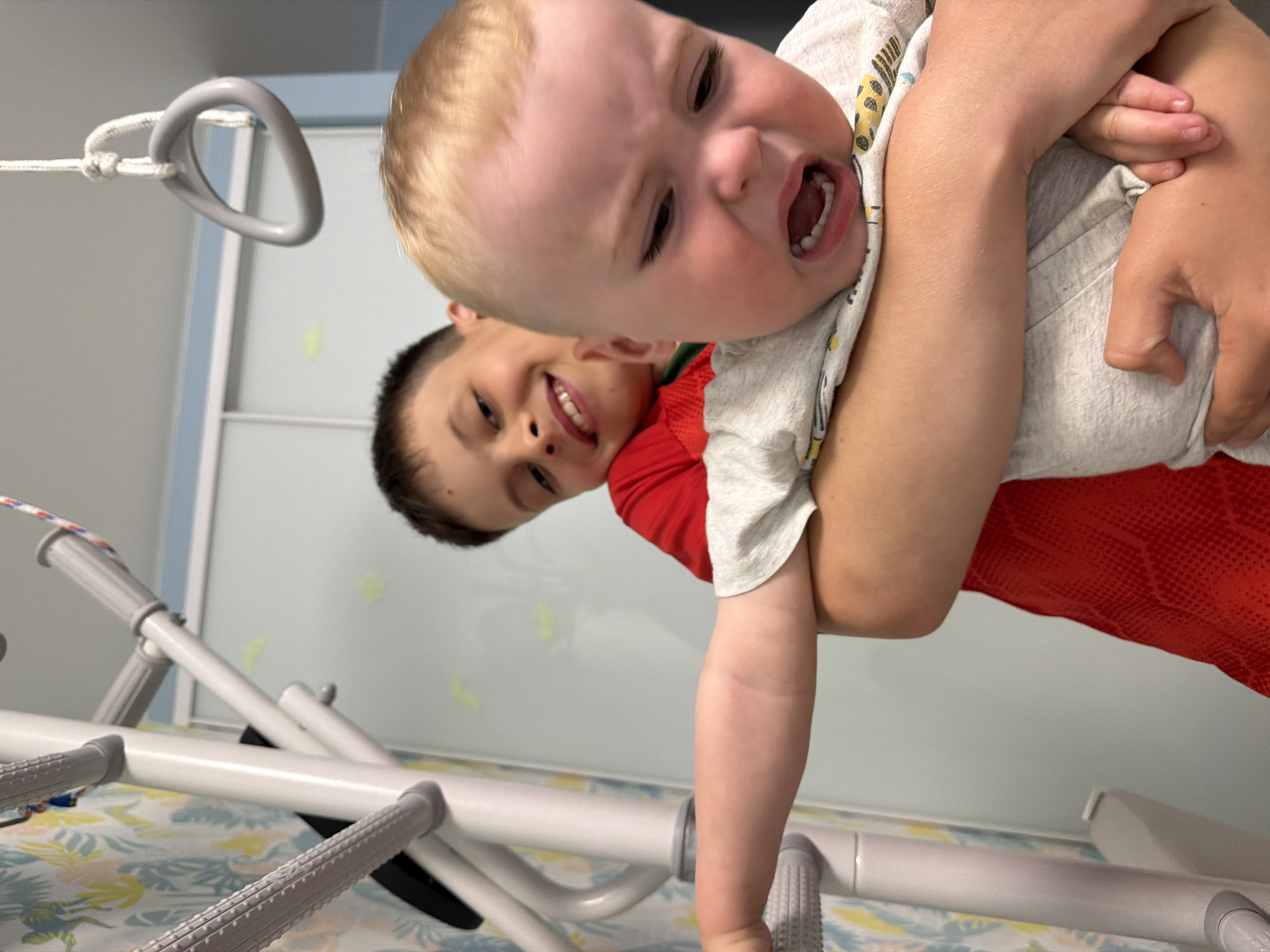 This image, taken in Votkinsk, Russia, captures a playful moment between two young boys in what appears to be an indoor setting. The photo is taken from a low angle, looking upwards, and is oriented at a 90-degree rotation.
In the foreground, a young boy with light blond hair and fair skin is being held by an older boy. The younger boy's face is prominent, with his mouth wide open, suggesting a shout of excitement or laughter during play. He is wearing a light-colored t-shirt with subtle yellow patterns. His arms are wrapped around the arm of the older boy, who is smiling broadly in the background, wearing a red t-shirt.
To the upper left, a gray, triangular D-ring is visible, secured by a white rope, hinting at a potential play structure or a therapy device. Across the lower part of the image, a white and gray support structure with textured grips runs horizontally, which could be part of a bed, a playpen, or medical/therapy equipment. The background features light-colored walls or partitions, with a faint floral pattern visible on what appears to be a sheet or wallpaper. The overall emotion conveyed is one of joy and active play between the two children. No visible text is present.