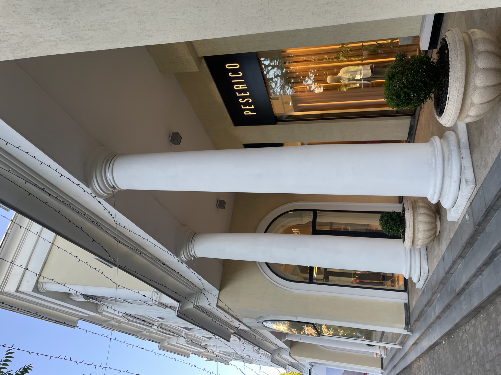 A storefront with the name "PESERICO" is visible above a display window. The window reflects trees and the sky, suggesting it might be daytime. Inside the store, a mannequin is dressed in a light-colored outfit. A small green bush in a decorative pot sits to the right of the storefront. The building features large white columns, ornate trim, and strings of small lights draped along the overhang. The ground in front of the store is paved with stone tiles. The scene appears to be a commercial street or plaza, possibly in a warm climate given the greenery and outdoor lighting. The overall impression is one of an upscale boutique or retail establishment.