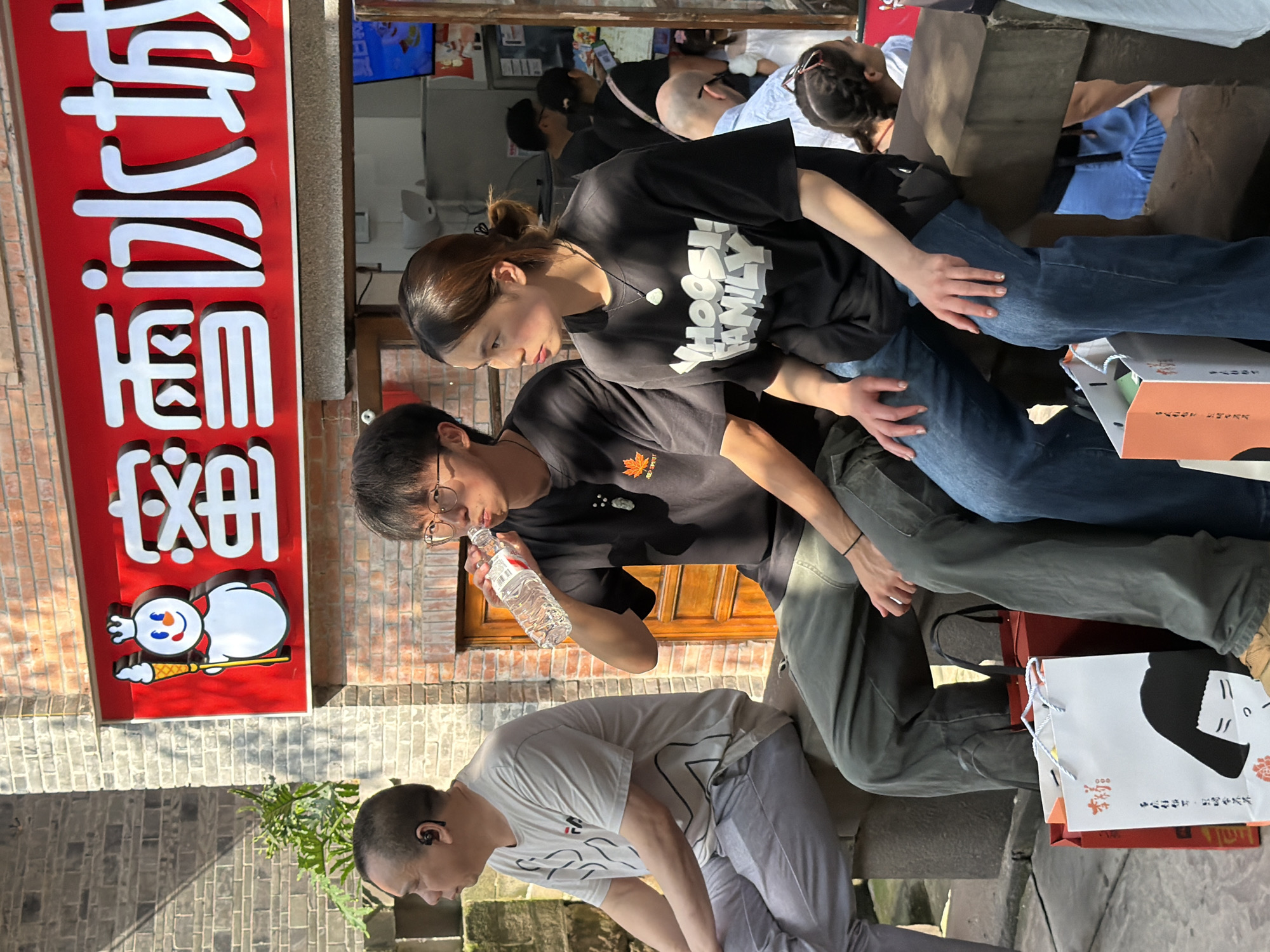 The image depicts a street scene in Yuzhong District, China, with a bright red sign featuring a snowman character and Chinese text that reads "Xue Bing Cheng" (Snow Ice City). In the foreground, three individuals are seated on a low stone wall. The person on the left, an adult male with short hair and wearing an earpiece, is dressed in a light grey t-shirt and grey pants. Next to him, a young male with glasses and dark hair is drinking from a bottle of water. He wears a black t-shirt with a small orange leaf graphic. To his right, a young female with brown hair tied back and wearing a black t-shirt with the text "Whoosh! Family" is seated. She has a pendant necklace. In the background, other people are visible, some queuing at a food establishment, and the building facade is made of red brick. Two shopping bags are placed on the ground in front of the seated individuals, one white with a graphic of a woman's face and the other red. The scene is illuminated by sunlight, casting shadows.