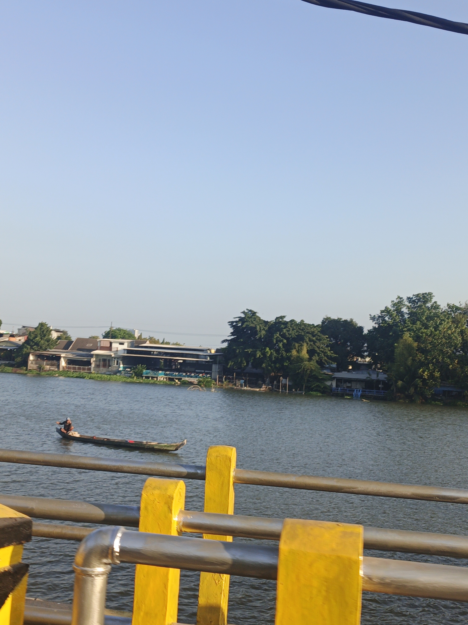 The image depicts a wide river with a clear sky above. In the distance, along the riverbank, a row of buildings and lush green trees are visible. One building appears to be a restaurant with outdoor seating. A lone person is seen rowing a long, narrow boat on the river. In the foreground, a metal railing with yellow painted supports runs horizontally across the frame, suggesting the vantage point is from a bridge or elevated walkway. The overall scene evokes a tranquil, everyday moment in a waterside community.