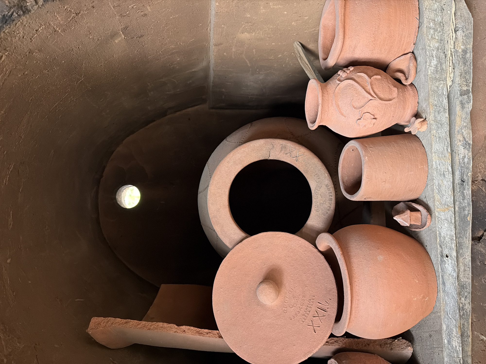 Inside a pottery kiln in Makatubani, Georgia, various unfired clay pots, lids, and shards are arranged. The interior of the kiln is a dark, cylindrical space, illuminated by a single point of light in the distance. Several terracotta-colored vessels are stacked and positioned near the kiln's opening. Some pieces are adorned with markings, possibly makers' stamps or serial numbers. A wooden plank is positioned next to the pottery.