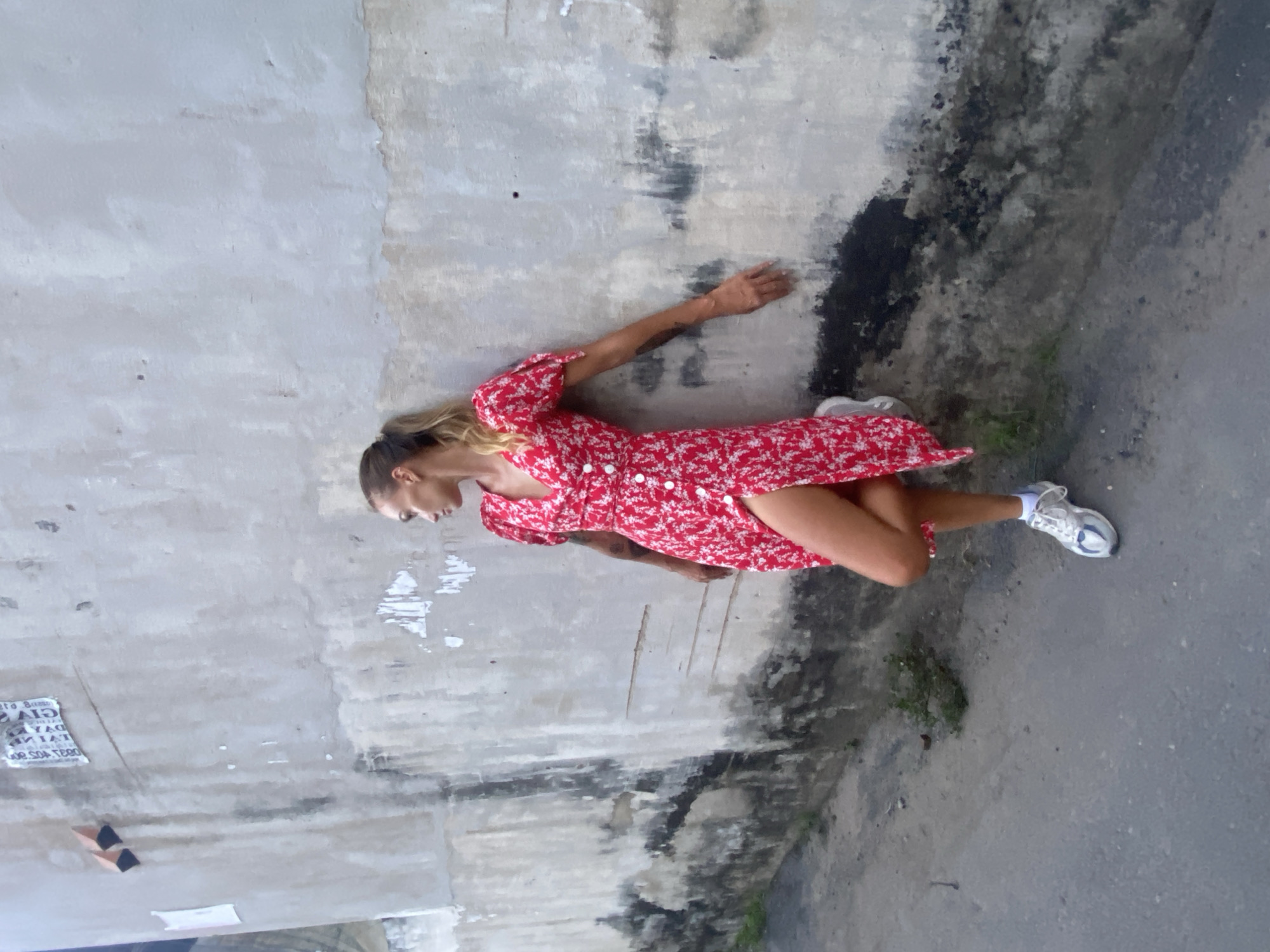 A woman lies on the ground, partially supported by a wall in an urban environment. She is wearing a short, red, floral-patterned dress and athletic shoes. The wall is grey and weathered, showing signs of age and damage. The ground is a mixture of concrete and dirt. A faded poster is affixed to the wall. The location is likely in or near Đồng Nai, Vietnam, given the prompt's reference.