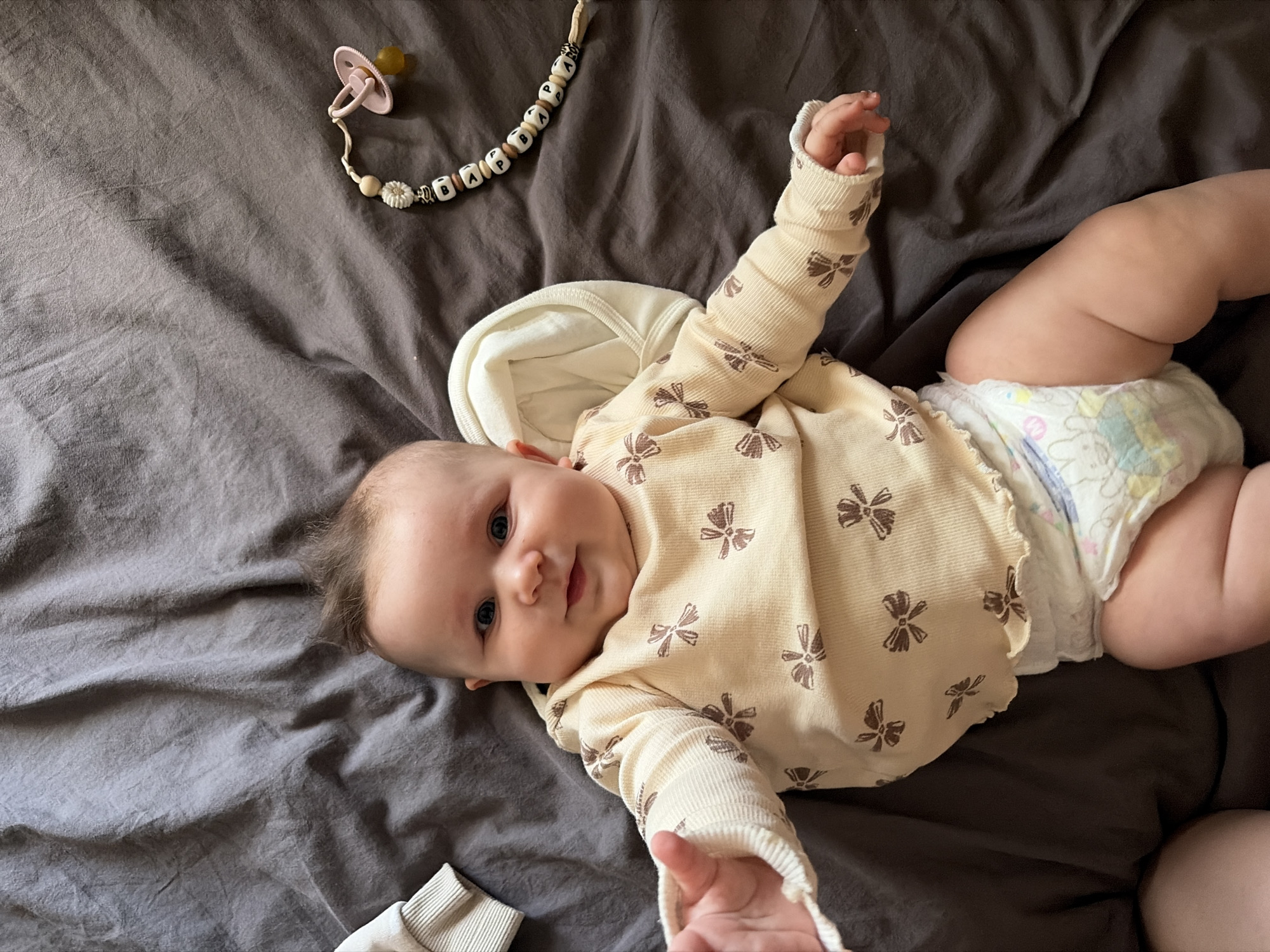 A baby is lying on a bed with a grey blanket. The baby is wearing a light yellow long-sleeved shirt with a pattern of brown bows and a diaper. The baby has blue eyes and dark hair that is styled in a mohawk. A pacifier on a beaded string is to the right of the baby. The image is a close-up shot, focusing on the baby's face and upper body. There are no discernible geographical landmarks or indications of the city of Chaikovsky, Russia.