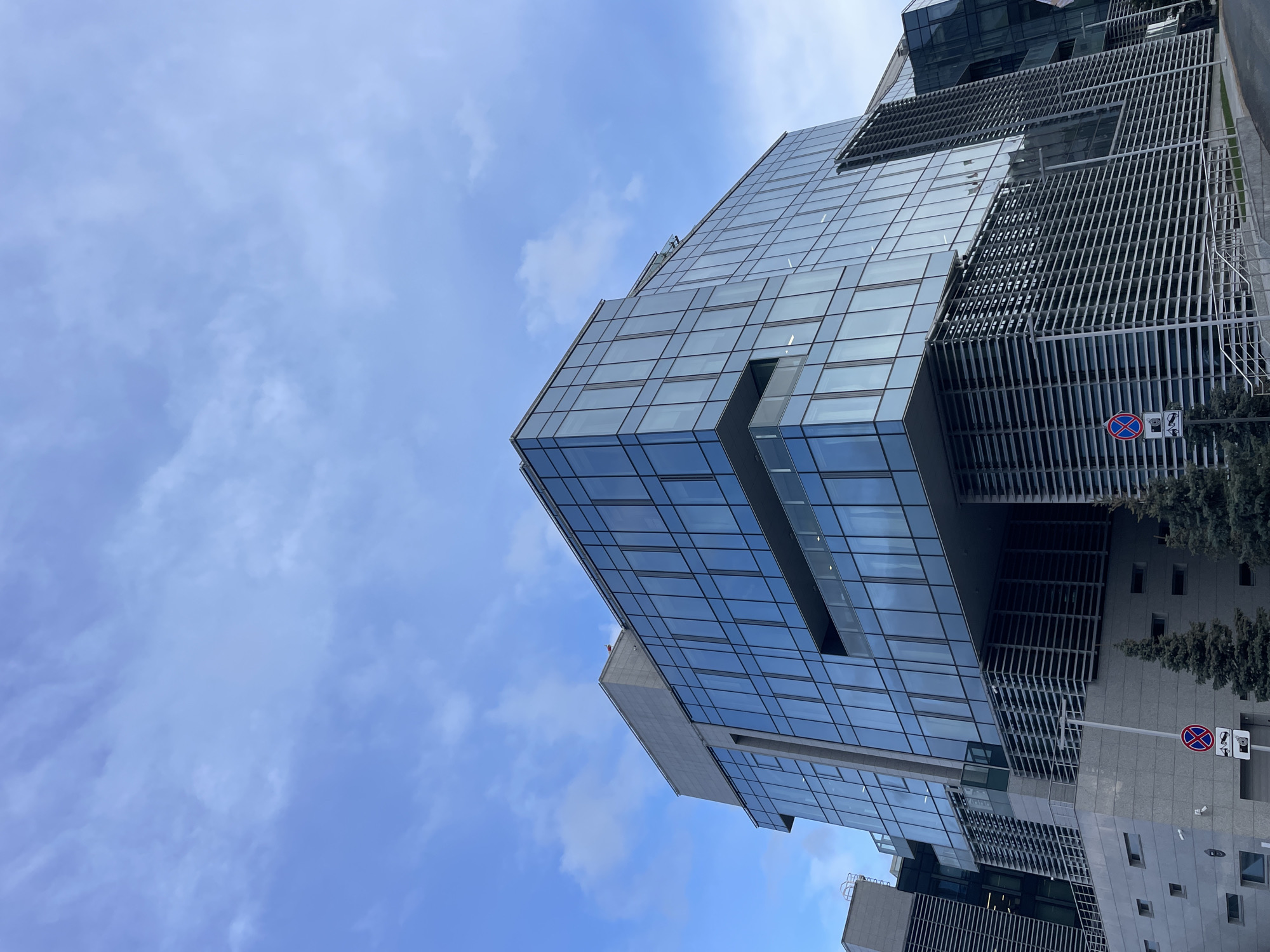 A modern, multi-faceted building is captured from a low-angle perspective, pointing upwards towards a blue sky dotted with scattered white clouds. The building features extensive reflective glass facades, which mirror the sky, and sections incorporating sleek, vertical metal slats or fins that contribute to a contemporary aesthetic. A portion of the building towards the lower right exhibits a grey, possibly concrete or stone, finish with several rectangular windows.
At the ground level on the right side of the image, green evergreen trees are visible. Two road signs are also present: a circular red and blue "no stopping" or "no waiting" sign, and a rectangular white sign below it displaying two black pictograms—one resembling a security camera and another, possibly indicating a specific restriction or zone.
The scene conveys an urban environment, likely a business or corporate area, during daylight hours on a partly cloudy day. There are no people or specific activities depicted, placing the focus on the building's architecture and its interaction with the sky.