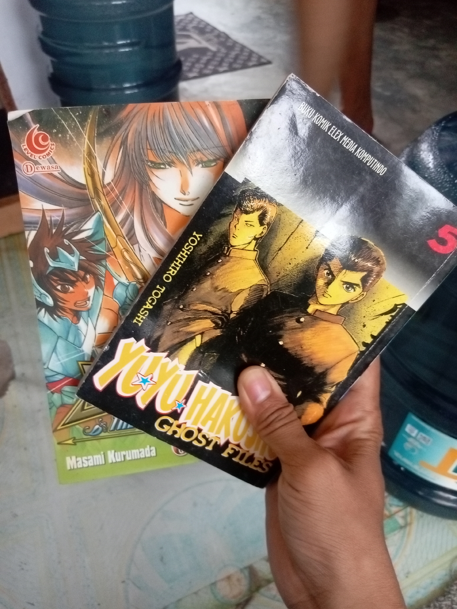 The image shows two comic books held by a person's hand, in what appears to be an indoor setting. One comic, titled "YUYU HAKUSHO GHOST FILES," is held at an angle, with a partial view of its cover featuring two animated male characters. The other comic, partially obscured, shows an anime-style female character with long hair and a blue armored figure. The background reveals a dark blue plastic container and a patterned surface. The text "LEVEL COMICS" and "Dewasa" is visible on the second comic. The text "BUKU KOMIK ELEX MEDIA KOMPUTINDO" and "YOSHIHIRO TOGASHI" are also present on the comic book covers. The overall lighting suggests an indoor environment with ambient light. The image does not provide specific geographic indicators to pinpoint a location within Batam, Indonesia.