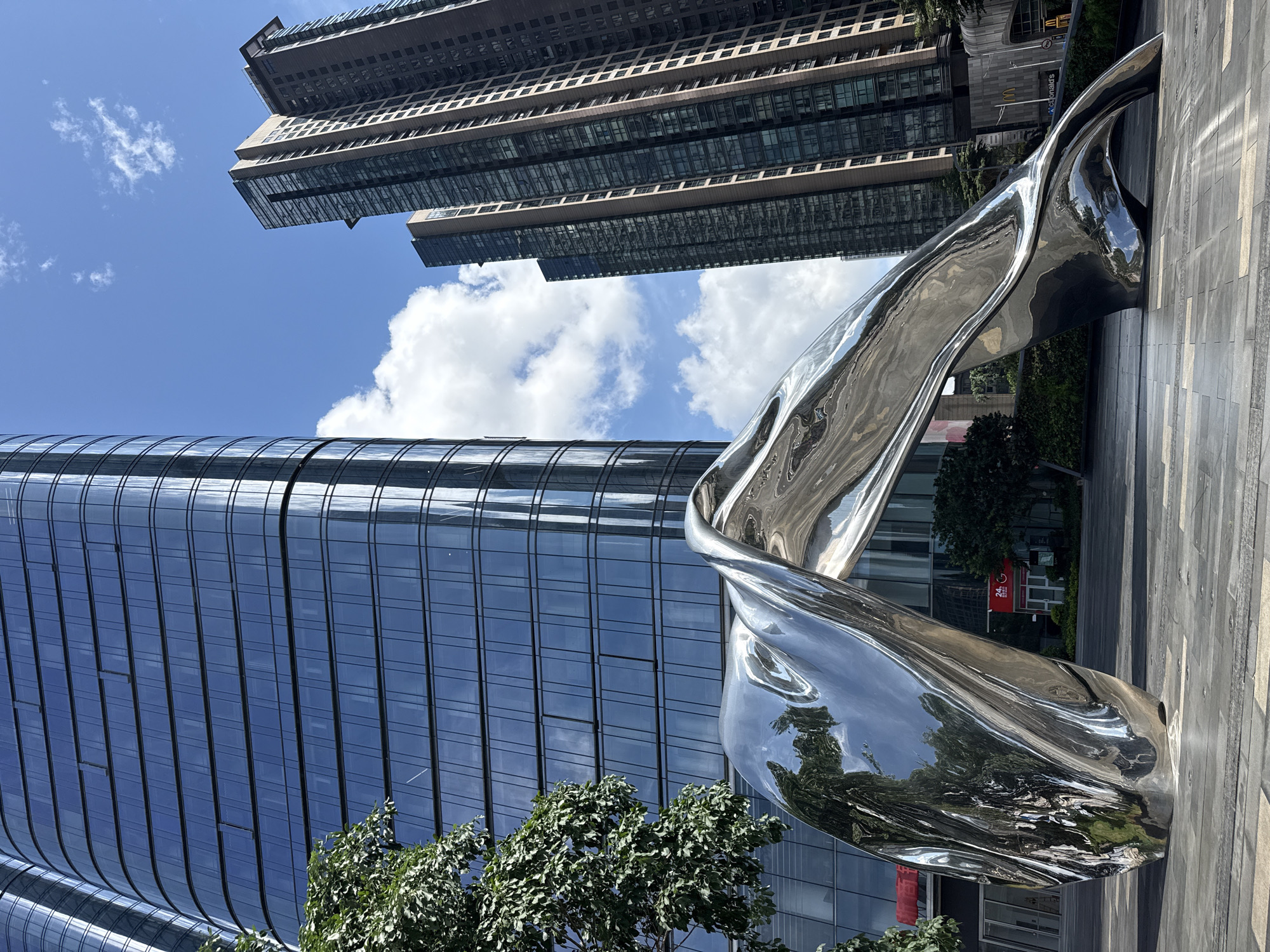 A low-angle, outdoor shot captures a twisted, shiny metal sculpture set against the backdrop of modern skyscrapers in Futian District, China. The sculpture’s reflective surface mirrors the surrounding buildings and sky, creating a distorted, abstract effect. A tree with green foliage is positioned in the lower left corner of the image, partially obscuring the base of the building behind it. The buildings are characterized by their glass facades, reflecting the blue sky and scattered white clouds. The skyscraper in the upper part of the image has a more traditional architectural style. The ground is paved with light-colored rectangular slabs. A red sign is visible between the sculpture's legs.