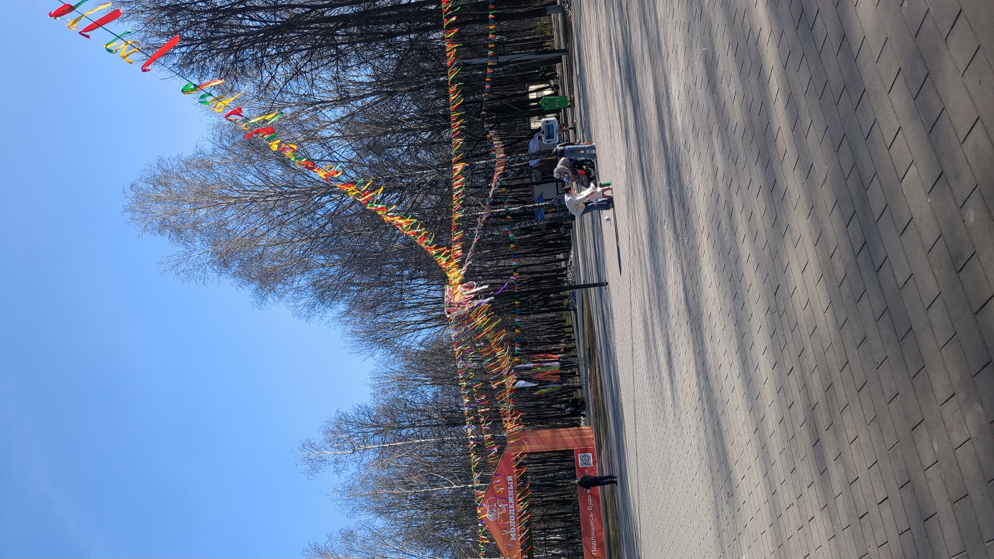 This media file depicts a wide, paved park pathway adorned with numerous colorful decorations under a clear blue sky. The path is flanked on both sides by a dense line of deciduous trees with bare branches, suggesting late winter or early spring.
Long strings of small, triangular flags and flowing ribbons, primarily in shades of red, green, and yellow, are strung between the trees, forming an extended canopy over the path. In the foreground, an orange inflatable archway spans the pathway. Visible text on the archway includes "11-Й МОЛОДЕЖНЫЙ" (11th Youth) and "ПОДПИШИСЬ. БУДЬ В" (Subscribe. Be in/on...). A QR code is also present on the archway.
Several people are visible in the scene. One person stands near the archway, while further down the path, a few individuals are seated on park benches or walking. A green trash can is also discernible near the benches.
The bright sunlight casts long, distinct shadows from the trees and overhead decorations onto the grey rectangular pavers of the path, indicating a clear and sunny day, likely late morning or early afternoon. The vibrant decorations and archway suggest a public event or celebration, possibly a youth event or race, taking place in a park in Ulyanovsk, Russia.