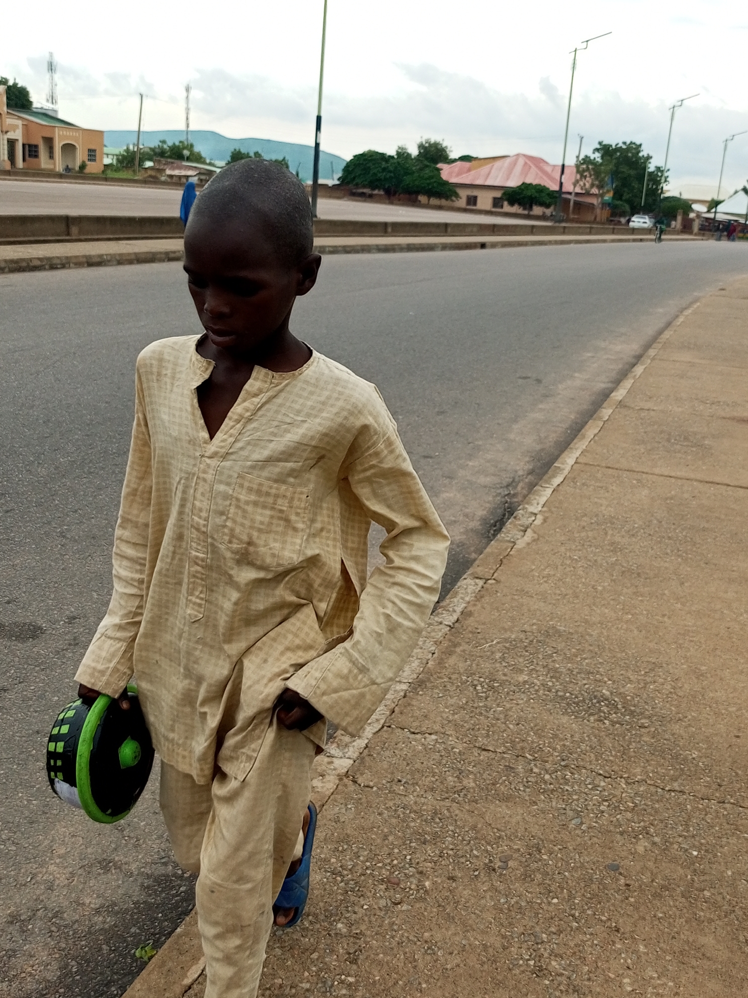 A young boy with dark skin and a shaved head walks along a cracked concrete sidewalk next to a paved road. He wears a light beige, checkered long-sleeved tunic with a V-neck and matching pants. In his left hand, he carries a round, black object with green accents, resembling a portable speaker or a container. His right hand is in his pocket. Blue sandals are visible on his feet. The road is empty of traffic in the immediate foreground, with a concrete barrier separating it from the sidewalk. In the background, buildings with red roofs and light-colored facades line the other side of the road, interspersed with trees. Several utility poles with lights are also visible. The sky is overcast. The scene appears to be in Pantame, Nigeria.