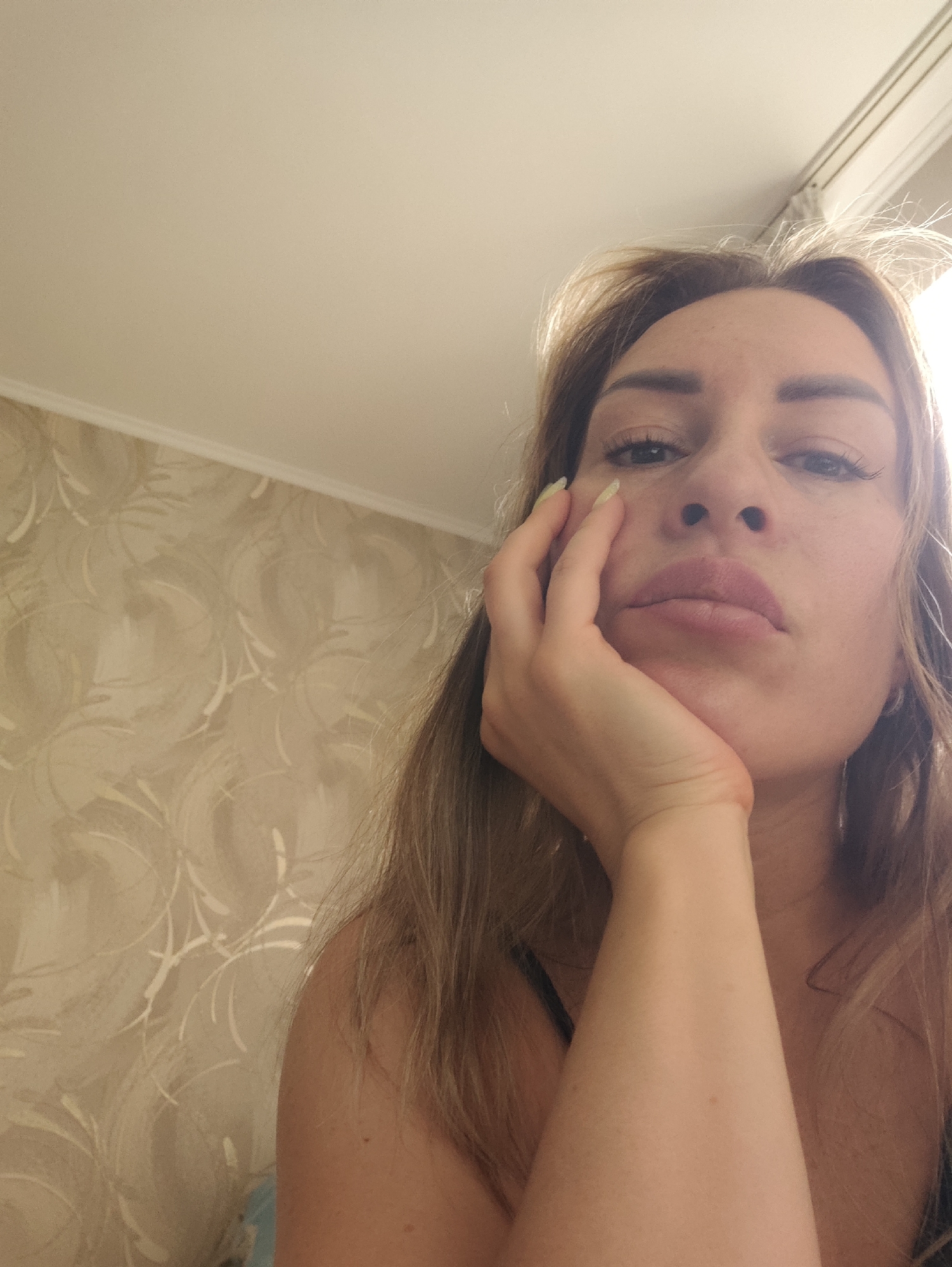 A close-up, eye-level selfie shows a woman with medium-length blonde hair, resting her chin on her hand. She is looking directly at the camera with a neutral expression. Her eyes are open, and her eyebrows are well-defined. Her lips are slightly parted. The background features patterned wallpaper in muted tones of beige and cream, and a section of a white ceiling with a molding. Sunlight is streaming in from the right side of the frame, creating a bright highlight on her hair and face. The image appears to be taken indoors, likely in a home setting, during daylight hours. No visible text is present. The overall mood is subdued and introspective.