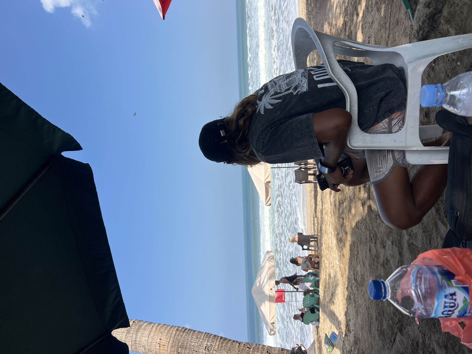A man wearing a black baseball cap and a dark t-shirt sits on a white plastic chair on a sandy beach, facing the ocean in Kuta, Indonesia. His t-shirt has a graphic print of a palm tree and buildings. He is holding a black object in his hands. In the foreground, a partially empty bottle of Aqua water sits on the sand next to a red plastic bag and a black backpack. The beach is lined with umbrellas and several people are seated on chairs under them. The ocean is visible with waves breaking, and the sky is a clear blue with a few scattered clouds. A palm tree trunk is visible on the left side of the frame.