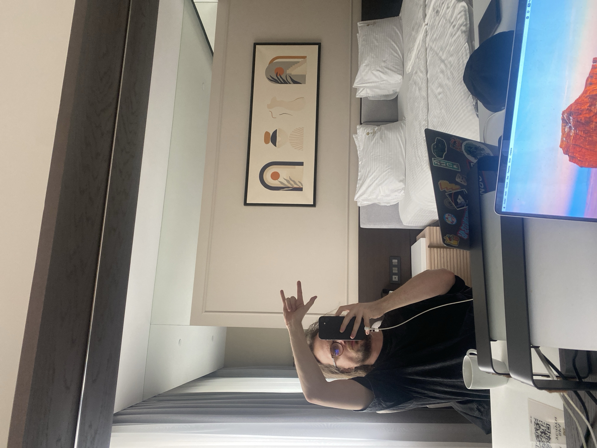A mirror selfie shows a man in glasses with medium length brown hair and a beard wearing a black shirt. He is holding a black phone and making the "rock on" hand gesture. The reflection shows a hotel room, including a bed with white linens, a framed piece of abstract art on the wall, a laptop with stickers, a large computer monitor, a white mug, and an access point label. The location is likely a hotel room but not verified to be in Elino, Russia.