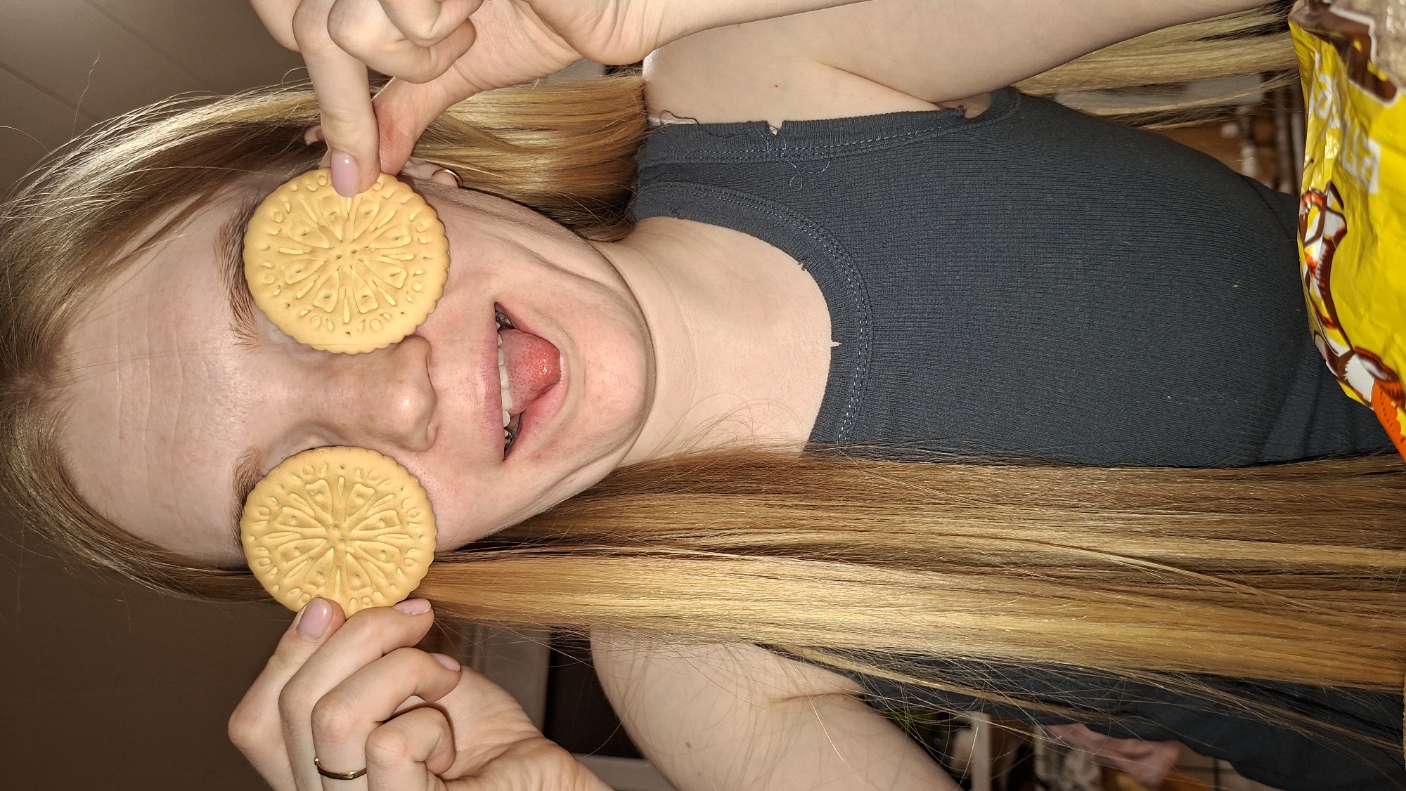 A woman is holding cookies over her eyes. She is wearing a gray tank top and has long, blonde hair. Her tongue is sticking out. There is a bag of chips behind her. The location cannot be determined.