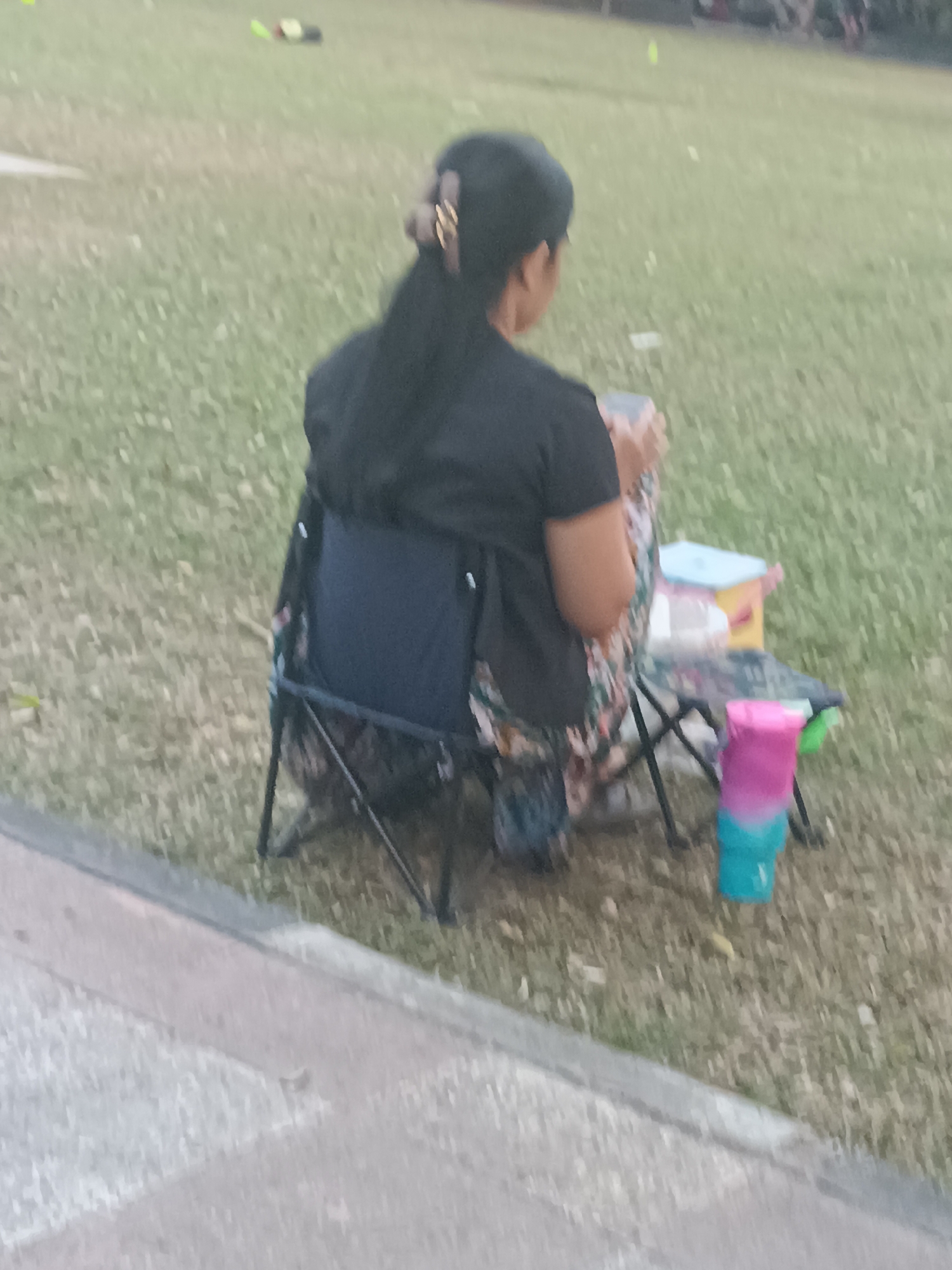 A woman with dark hair pulled back sits on a dark blue folding chair in a grassy area. She is wearing a black t-shirt and a floral patterned skirt. In front of her, on a small folding table, are several items including what appears to be a blue container, a yellow and pink container, and a multi-colored cup. The ground is covered in dry grass. A paved pathway is visible in the foreground. In the background, other people and some yellow objects are faintly visible on the grass. The overall lighting suggests it is either early morning or late afternoon. The scene appears to be a park or open recreational area within Sleman, Indonesia.