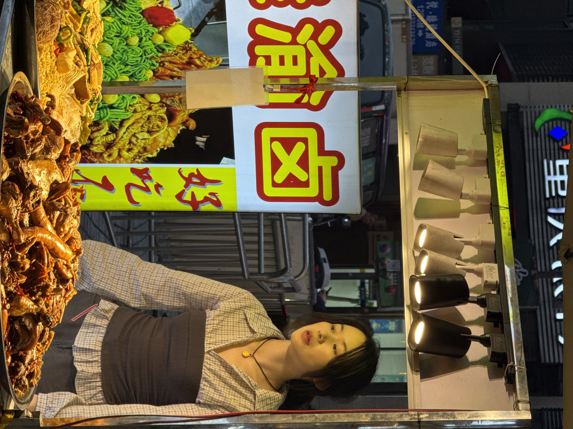 The image shows a food vendor, likely in the Yuzhong District of China. A woman in a gray plaid shirt and black apron stands behind a food cart. The cart displays a variety of food items, including a large tray of braised meats and a colorful poster showing different noodle dishes. Above the food, a sign in Chinese characters is visible. The cart is lit by a row of small spotlights. The background shows a typical urban street scene, with cars and shops.