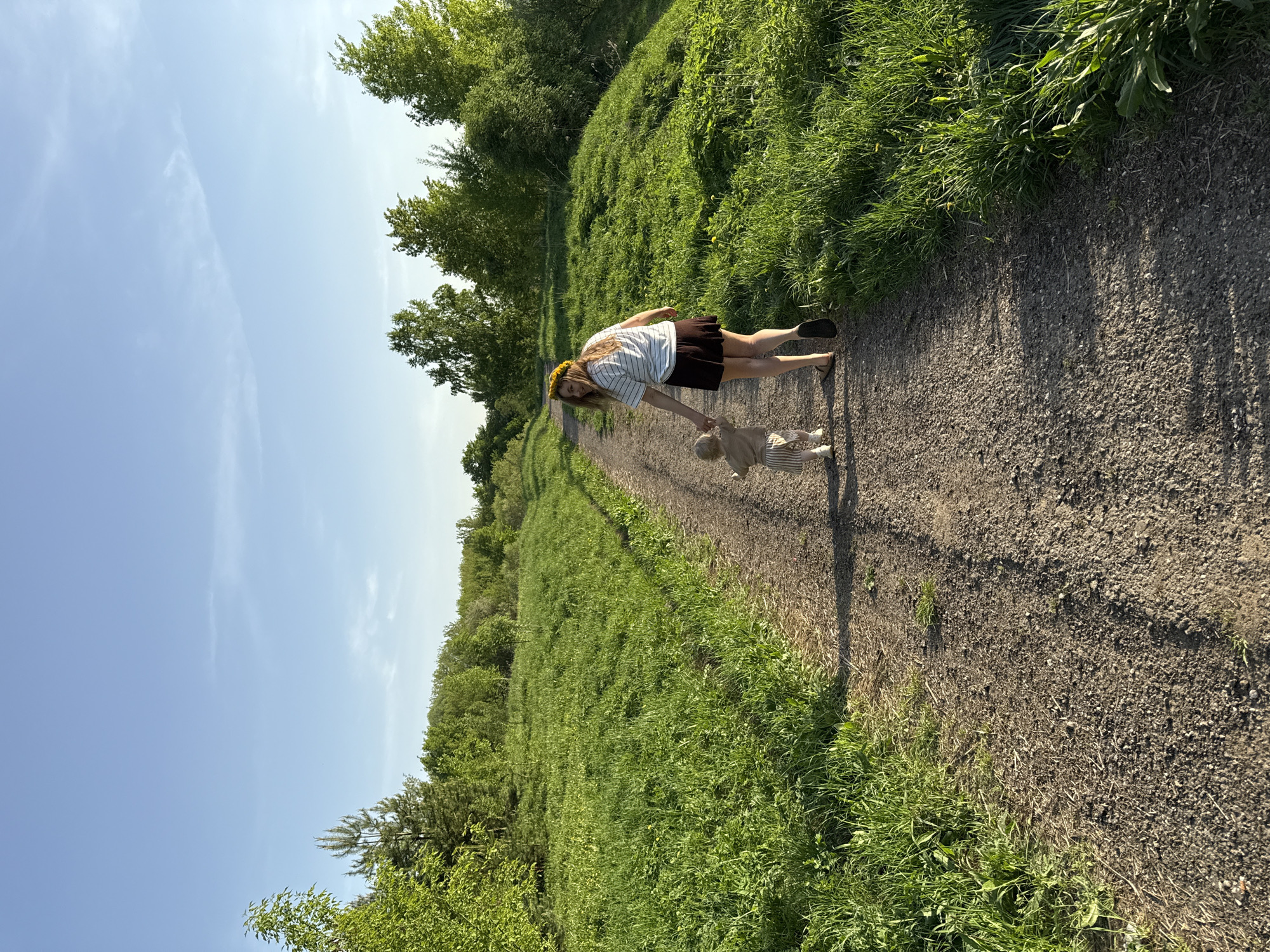 The image depicts a woman and a young child walking on a dirt path in a green, outdoor area, possibly a park or field. The woman, wearing a striped shirt, brown skirt, and what appears to be a floral headband, is holding the child's hand. The child is wearing a striped outfit. The path is surrounded by green vegetation and trees, and a clear blue sky is visible in the background. The location is not discernible from the image.