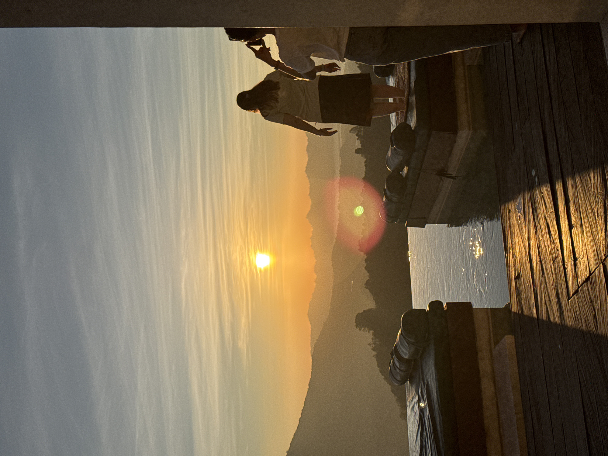 This image captures a sunrise or sunset over the mountainous landscape of Kintamani, Indonesia, viewed from a high vantage point, likely a resort or viewpoint terrace. The sky transitions from light grey-blue at the top to vibrant golden-orange and yellow closer to the horizon, with faint, streaky clouds. The sun, a bright, intense orb, is positioned slightly left of the image's vertical center, casting strong golden light across the scene and producing a prominent circular lens flare to the left of the primary subject.
Multiple layers of mountains are visible in the background, appearing progressively hazier and more silhouetted in shades of grey-green with increasing distance. In the midground, an infinity pool extends across the width of the frame, its surface reflecting the warm sky and sparkling with sunlight.
Two lounge platforms, each topped with dark, cylindrical cushions, are positioned at the edge of the pool. On the right-side platform, a person with long, dark hair, wearing a light short-sleeved top and a dark skirt, stands barefoot, facing away from the camera and looking out at the mountain view. Their arms are slightly extended downwards. To the immediate right, partially obscured by a grey vertical architectural element, another individual in a light-colored short-sleeved top and dark trousers is visible, holding a smartphone raised to capture a photograph or video of the panorama. The immediate foreground features a wooden deck, strongly illuminated by the low sun, highlighting its grain and texture.
