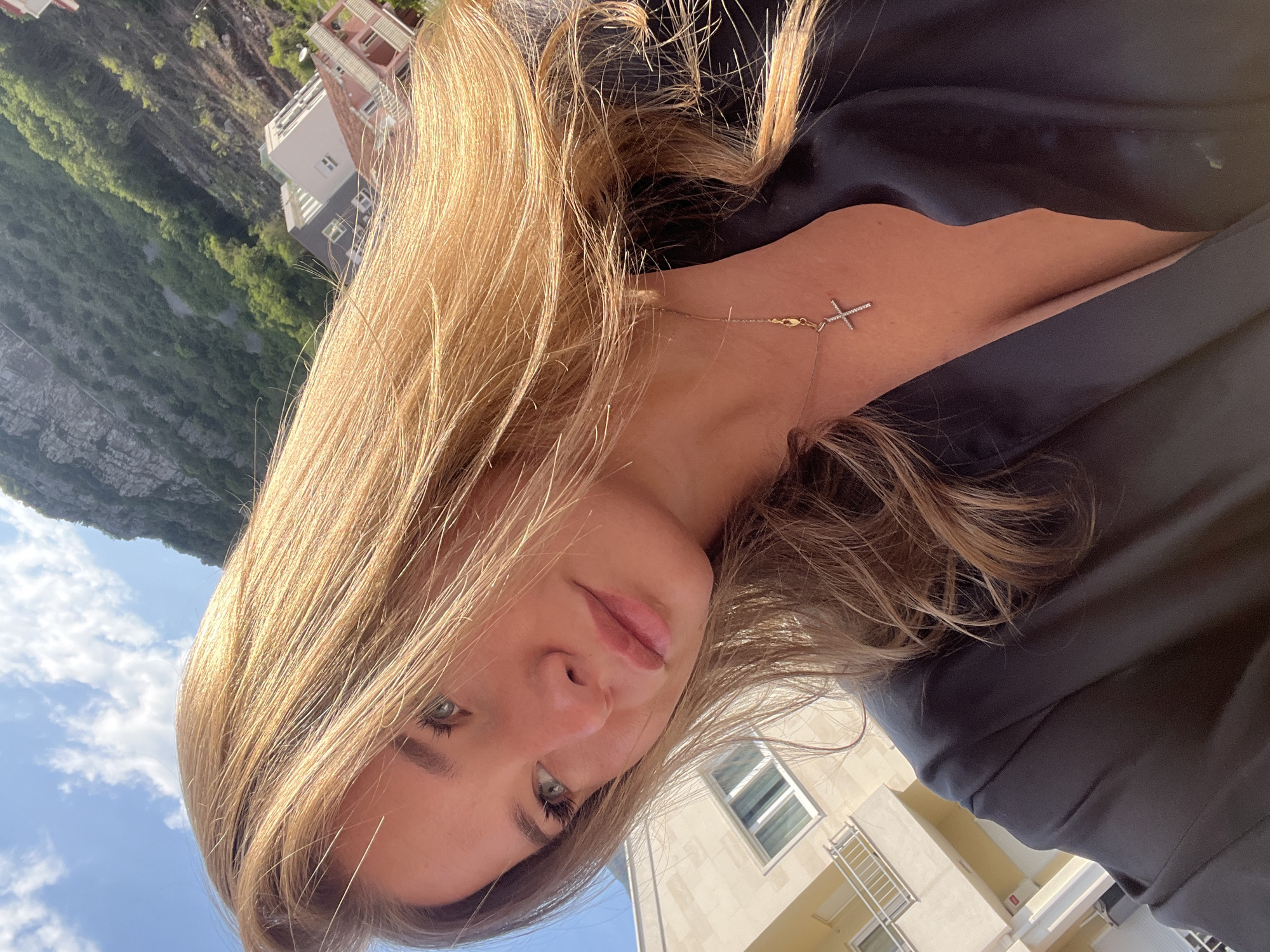 A young woman is captured in a close-up selfie, taken outdoors in bright daylight. Her head is tilted, and the image itself is rotated, giving a dynamic, upward-looking perspective. She has long, light brown or blonde hair, styled to fall around her face and shoulders, catching the sunlight with significant highlights. Her eyes are light-colored, possibly blue or green, and she has full lips with a subtle, natural expression. She wears a simple black top or dress, and a delicate, gold-toned chain necklace with a small, silver-toned cross pendant adorned with tiny stones is visible on her décolletage.
The background reveals an outdoor setting. To her upper left (in the original orientation), a rugged, rocky mountain slope covered with green vegetation is visible, suggesting a natural, possibly coastal, landscape. Further down and to her right, modern buildings with windows and balconies can be seen. The sky is bright blue with scattered white clouds. The strong sunlight indicates it is daytime. Given the context of Budva, Montenegro, the mountainous terrain and buildings are consistent with the region's coastal and urban environment. No visible text is present in the image.