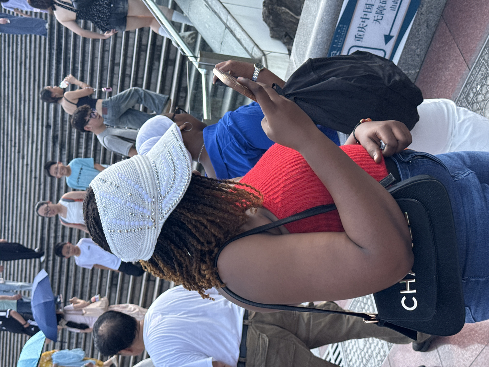 A medium shot captures a Black woman wearing a red ribbed tank top, blue jeans, and a white visor adorned with rhinestones. She is holding a black handbag with a gold "CHANEL" logo and looking down at her phone, which she is holding in her right hand. Her left hand is resting on her hip. Behind her, a person in a blue shirt is also looking at their phone, and further back, a group of people are ascending a wide, stone staircase. The scene appears to be in Yuzhong District, China, indicated by a sign in the background with Chinese characters and an arrow pointing left, which reads "重庆中国三峡博物馆" (Chongqing China Three Gorges Museum) and "无障碍通道" (Accessible passage). The environment consists of a paved walkway and stone steps, with a glass railing on the right.