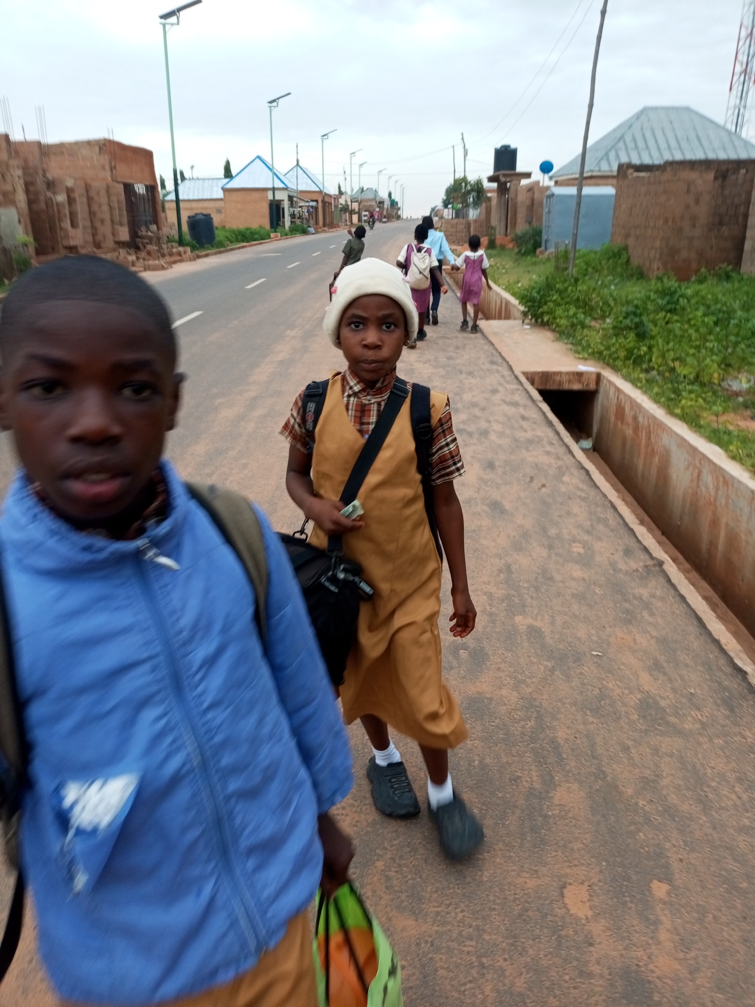 The image captures a street scene in Gombe, Nigeria, likely on a cloudy day. In the foreground, two children are walking on a paved road. The child on the left is closer to the camera, wearing a blue jacket and a backpack, with their face partially visible. The child on the right is walking ahead, wearing a tan uniform with a plaid shirt underneath, a white head covering, and carrying a bag. They are both wearing dark shoes and white socks.
In the background, several other people, including children in uniforms and adults, are walking on the same road. The street is lined with buildings constructed from brick and concrete, some appearing to be under construction. Solar-powered streetlights are visible along the road. A concrete drainage channel runs alongside the paved walkway. The overall impression is a typical urban environment in Gombe, with everyday activities taking place on the street.