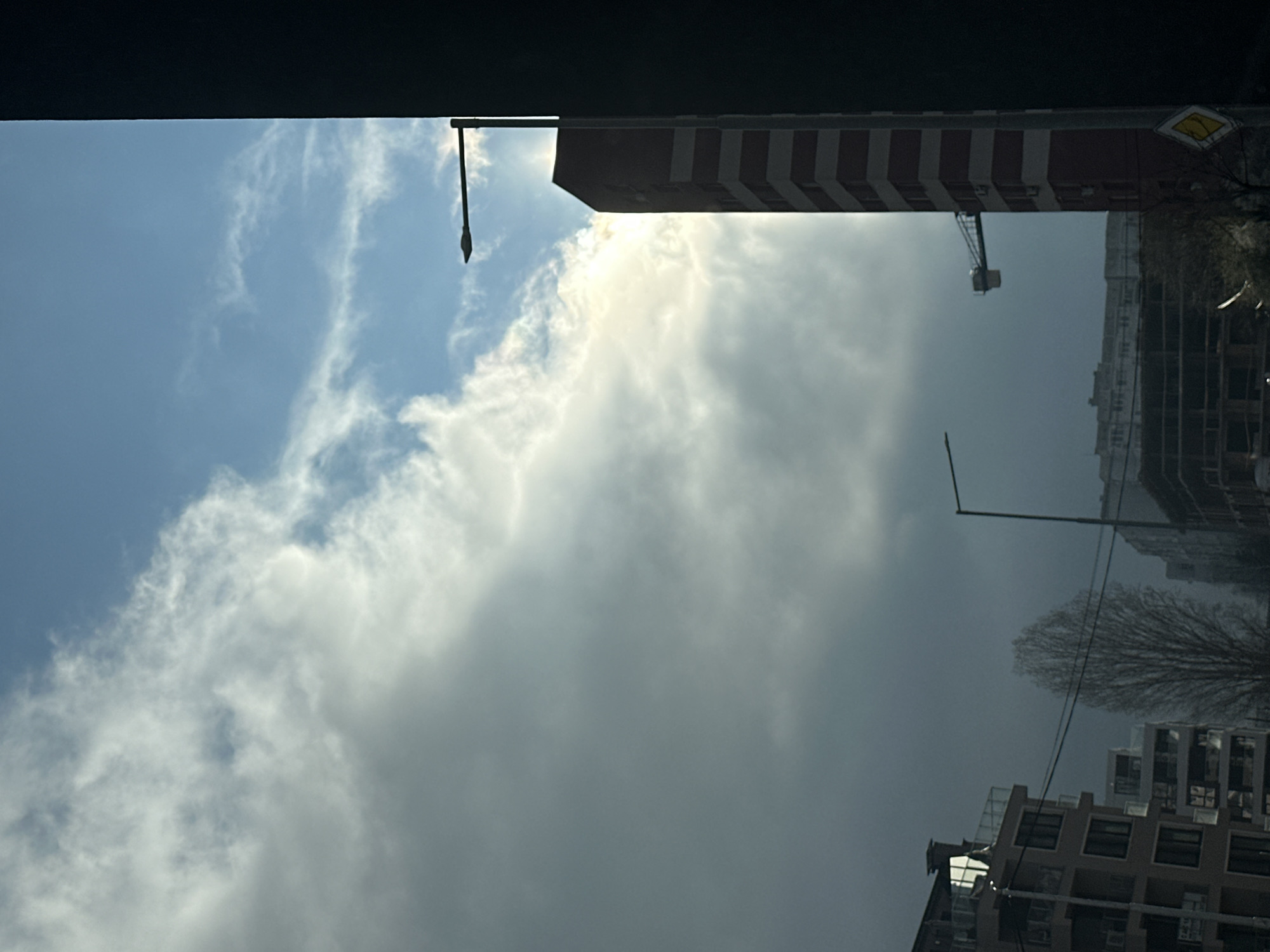 The image captures a view in Sofia, Bulgaria, looking upwards towards the sky. The sky is a mix of blue and white clouds, with some darker grey clouds obscuring parts of the view. In the foreground, a dark, indeterminate structure partially frames the top of the image. Several buildings are visible: one has a red and white striped pattern; a crane is positioned near a construction site. Trees and street lights are visible along with power lines.