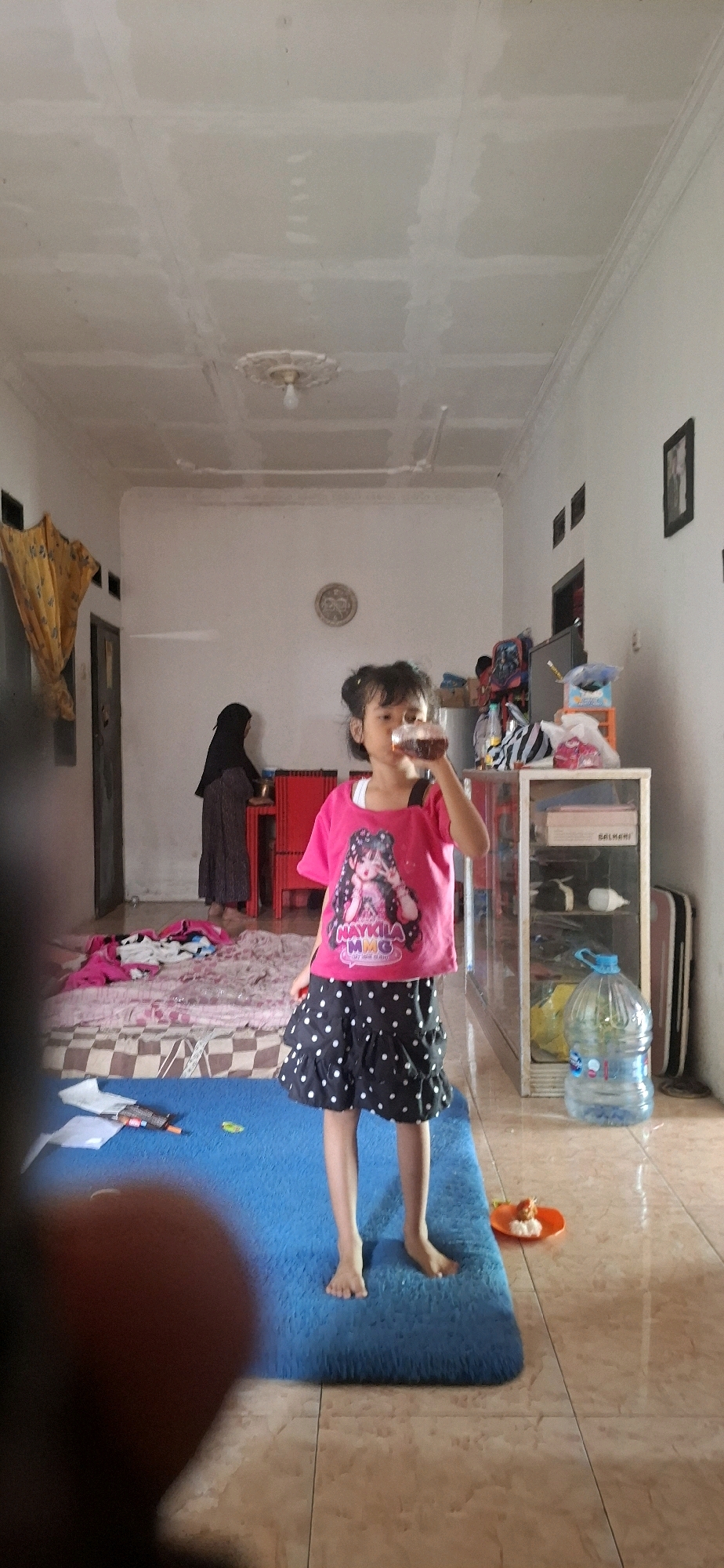 In Cibinong Hilir, Indonesia, a young girl with dark hair tied up in pigtails is standing on a blue rug, drinking from a bottle. She is wearing a pink t-shirt with a graphic of a cartoon character and the text "NAYKILA MMG" on it, and a black skirt with white polka dots. The room appears to be a living area with tiled flooring. To the left, a woman in a dark headscarf and dress is visible near a doorway. Behind the girl, there is a white cabinet with shelves containing various items, including a large water jug. The ceiling is white and shows evidence of construction or repair. The overall impression is of a casual indoor setting.