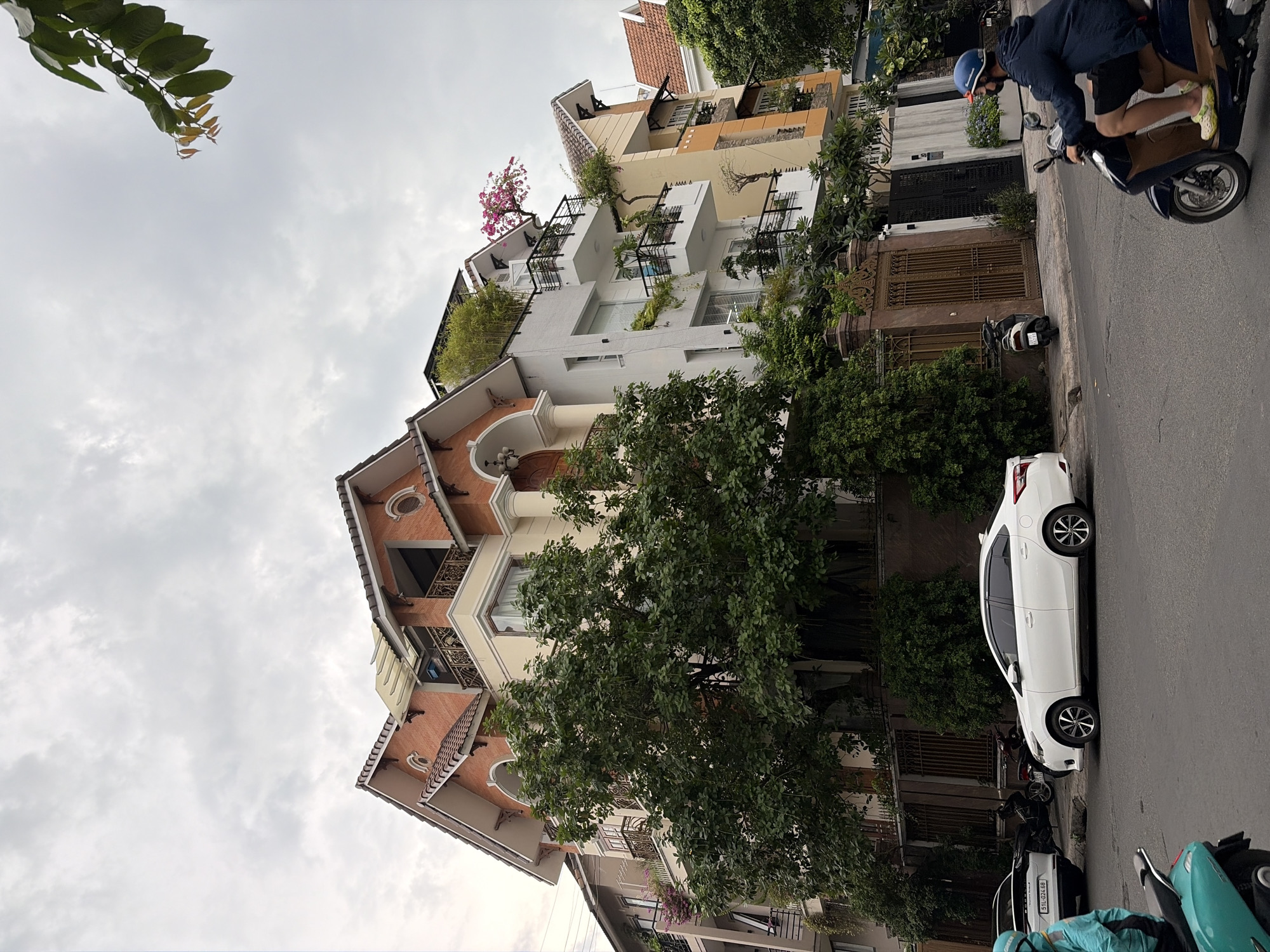 In Thủ Đức city, Vietnam, a white sedan is parked on the right side of a paved street. A person wearing a blue jacket, helmet, and shorts rides a dark-colored scooter from right to left in the foreground, passing by a large, leafy green tree. Behind the tree, an ornate, wrought-iron gate is visible. To the right of the gate, a multi-story building with white and yellow facades, balconies, and a red-tiled roof is situated. A pink flowering plant is visible on one of the balconies. Further to the left, another white sedan is parked, and a person in a turquoise jacket is partially visible on a turquoise scooter. The sky above is overcast.
