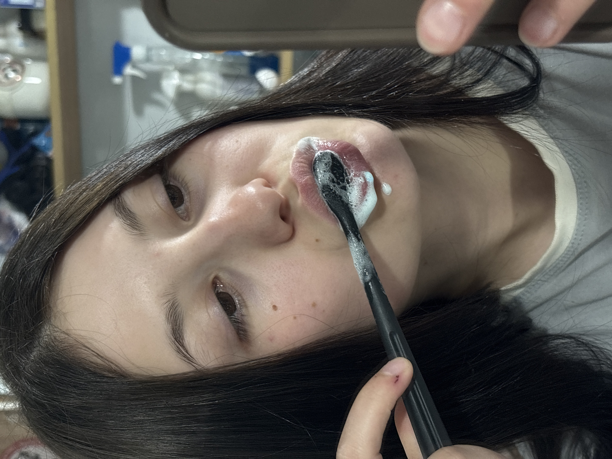A person is brushing their teeth with a black toothbrush and white toothpaste. The person has dark hair and light skin, and they are wearing a gray shirt. A shelf with various objects is visible in the background. Фоки, Russia is not apparent in the image.