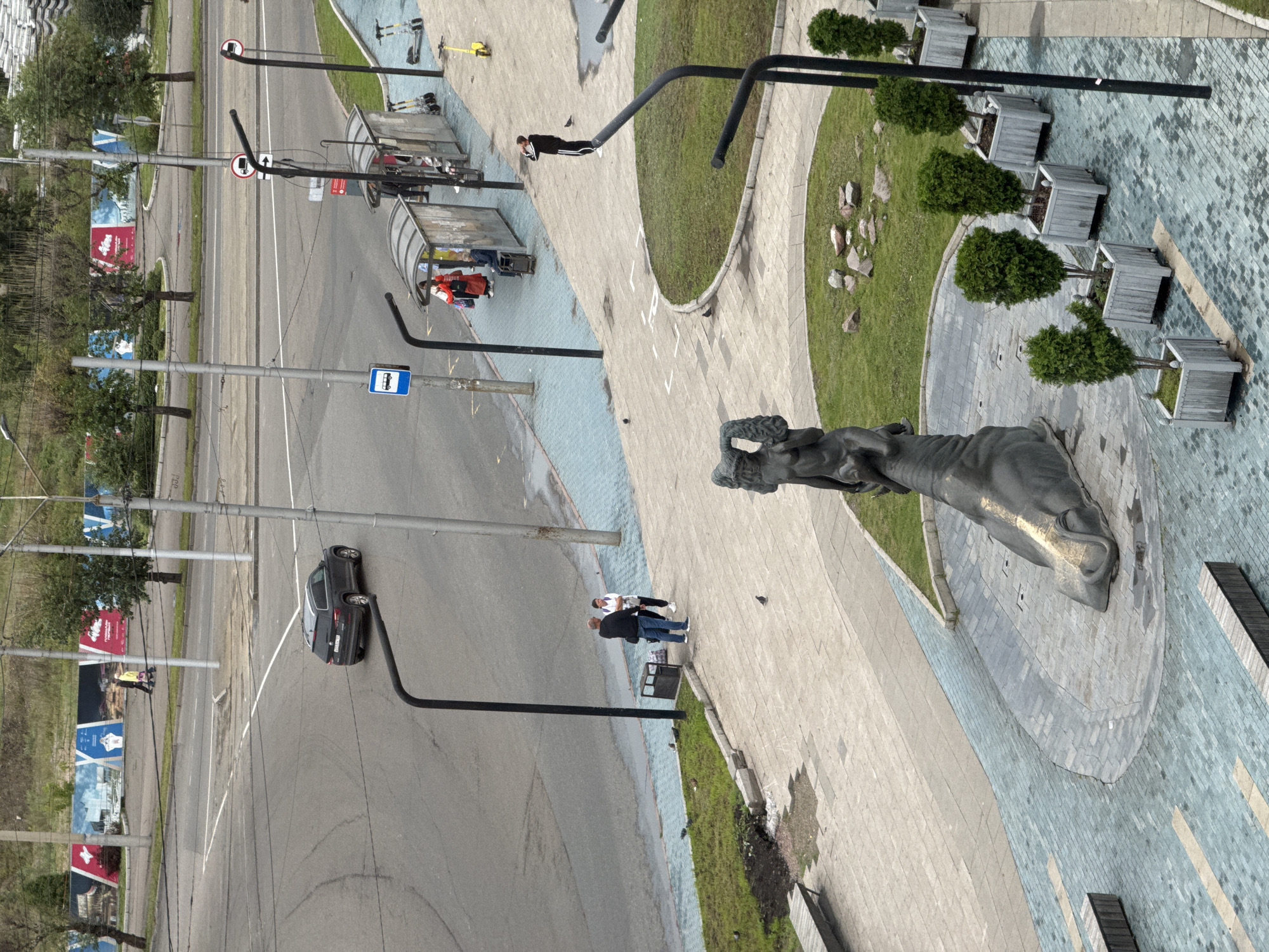 The image captures an urban scene in Krasnoyarsk, Russia, featuring a street intersection, a bus stop, and a public plaza with a statue. In the background, a dark SUV drives through the intersection, while people gather at the bus stop. The plaza is adorned with planters containing trees and a large bronze sculpture of a mythological creature. The ground is paved with textured tiles, and benches are strategically placed for seating. The overall impression is one of a lively, contemporary urban space.