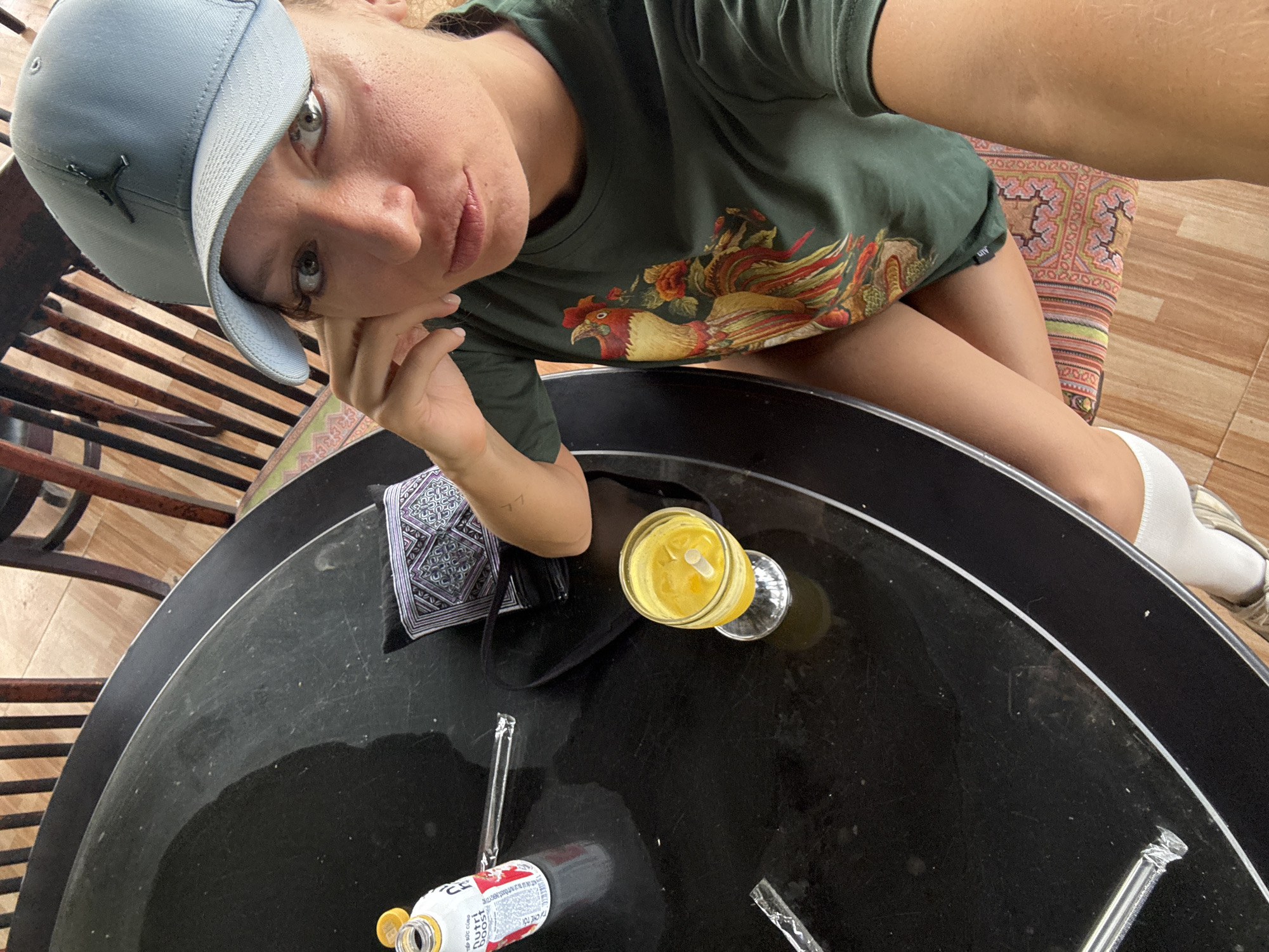 A person with light skin and light eyes, wearing a blue baseball cap and a green t-shirt with a rooster design, is looking at the camera. They are sitting at a round black table with a drink in a glass, a bottle of Nutri Boost, and discarded straws. A decorative cloth is on the table as well. The floor and chair suggest an indoor setting in Cốc Pài, Vietnam.