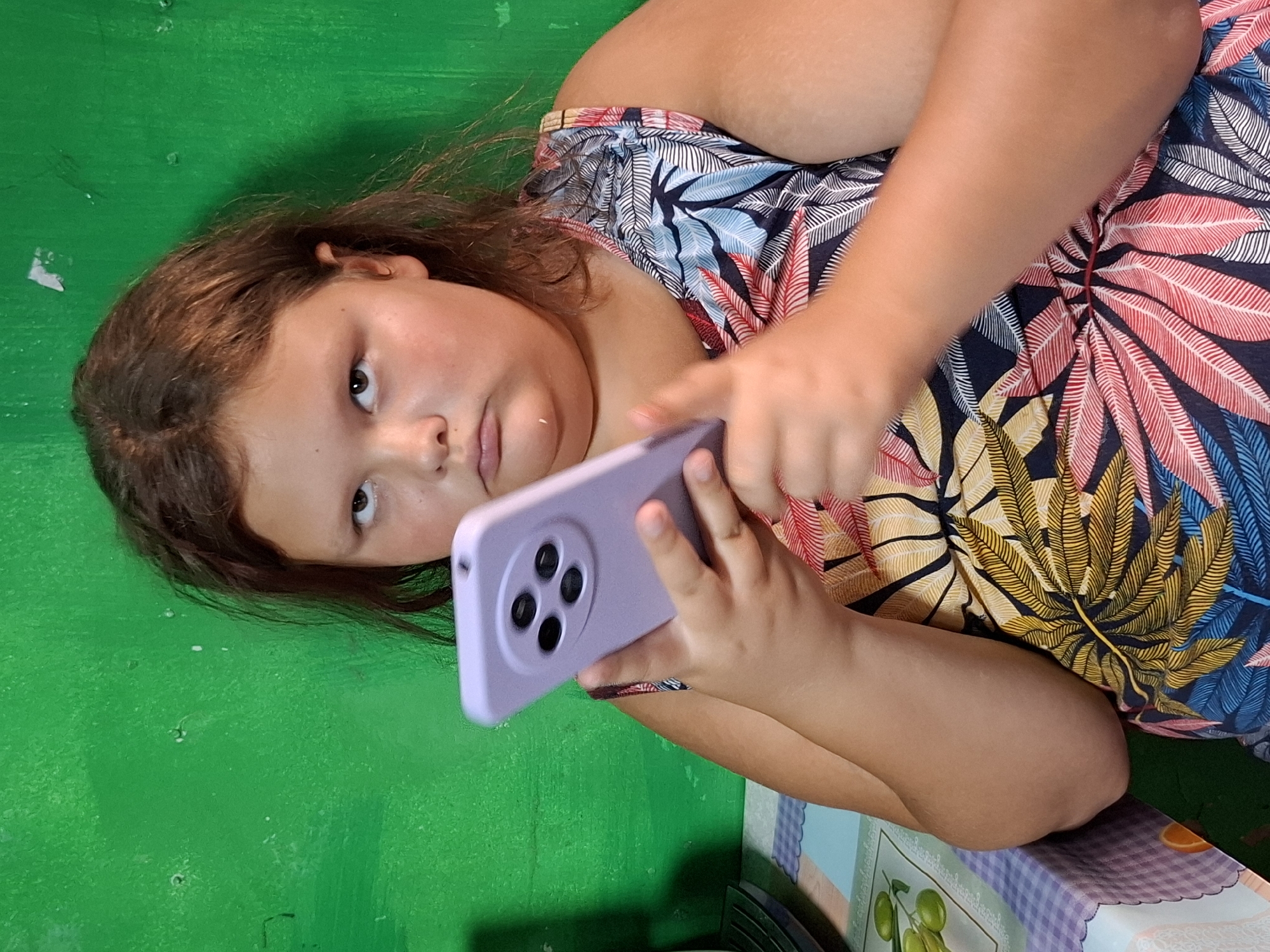A young girl with brown hair lies on a bright green surface, looking up at a purple smartphone she is holding with both hands. Her eyes are dark and appear curious. She wears a colorful, patterned top with large leaf designs in shades of pink, blue, and yellow. Her arms are bent as she holds the phone, with her left hand supporting it and her right index finger touching the screen. The phone case is a light purple and features a circular camera module with four black lenses. The background is a textured green surface, possibly a table or wall. A corner of a patterned surface, featuring a checkered design and fruit imagery, is visible on the right side of the image. The lighting suggests it is daytime, and the overall scene appears casual and domestic. There is no visible text in the image.