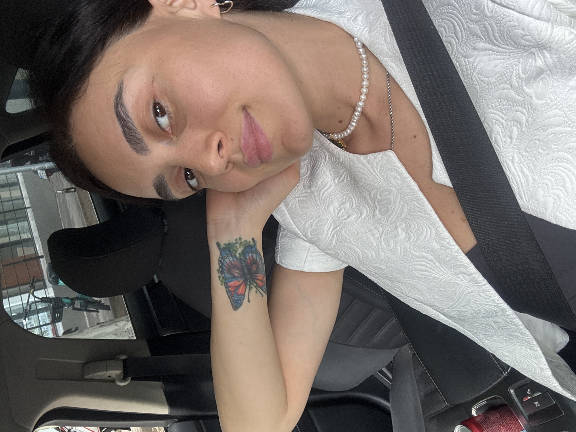 A woman with dark hair, wearing a white textured jacket and a pearl necklace, is seated in the driver's seat of a car. She has a butterfly tattoo on her left arm. The car appears to be parked or stopped, with a red soda can and car controls visible in the foreground. Outside the car window, a bicycle with a basket and a building facade can be seen. The image was taken in Tbilisi, Georgia.