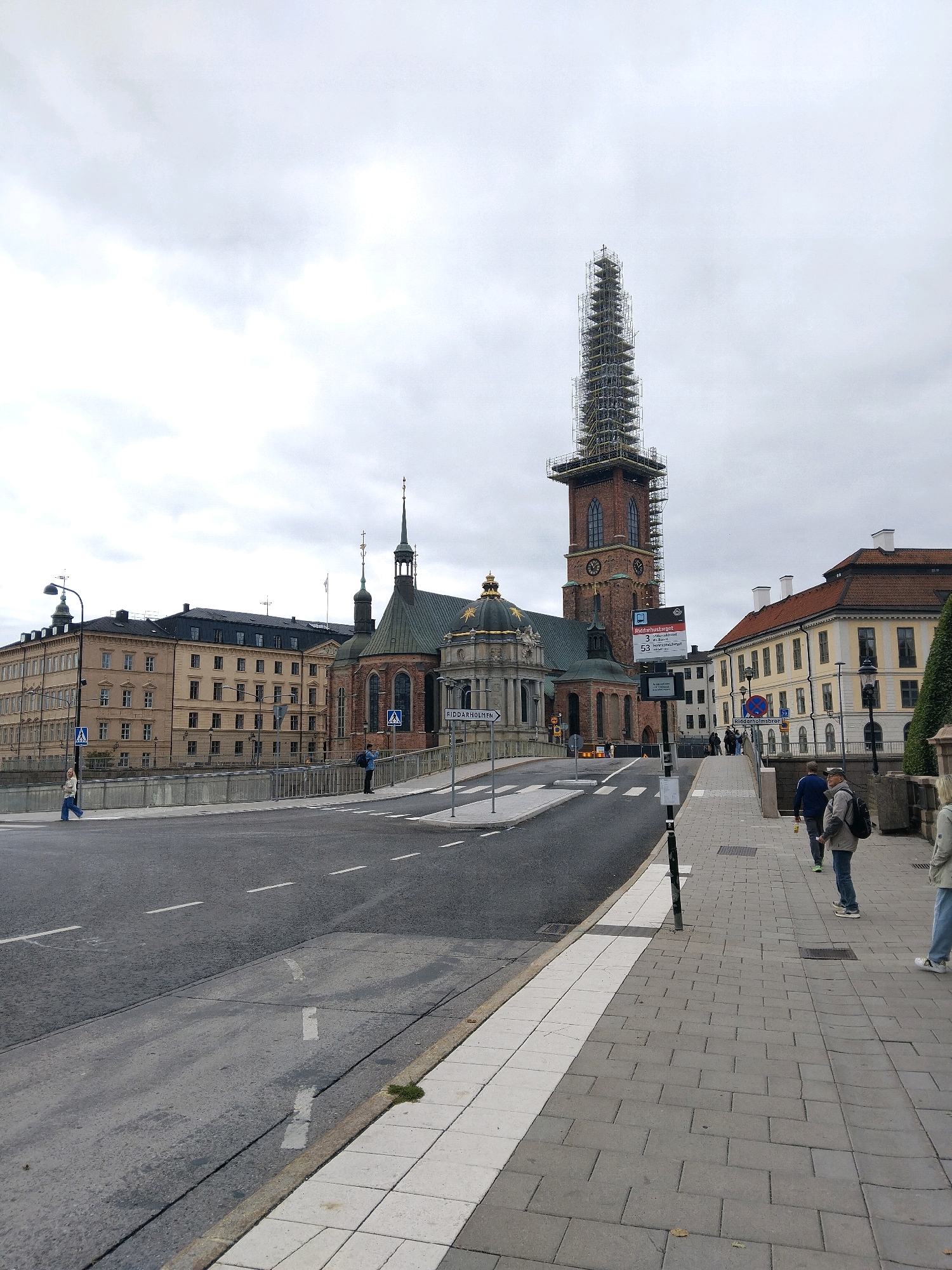 The image depicts a view of Riddarholmen Church in Stockholm, Sweden, on a cloudy day. The church's tower is under renovation and is covered in scaffolding. In the foreground, a street with a pedestrian crossing is visible, with a sidewalk to the right where several people are walking. The street is lined with buildings on the left and right sides of the frame, some of which appear to be historical. A lamppost is positioned on the sidewalk on the left side of the street. The sky is overcast, suggesting a cool or damp day.