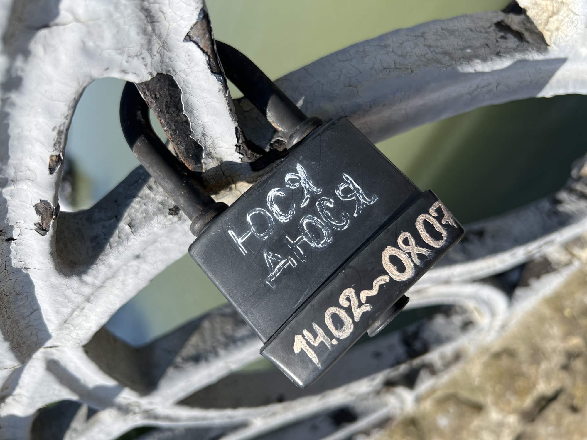 A close-up shot shows a black padlock attached to a white, ornate metal structure. The lock is inscribed with white lettering, reading "ЮСЯ ЛЮСЯ" on the main body and "14.02~08.07" on the lower part. The metal structure has chipped and cracked paint, revealing underlying rust. The background appears to be a body of water with a light green hue. This image likely depicts a "love lock" placed on a bridge. While not explicitly stated, the location of the image is in the city of Krasnodar, Russia.
