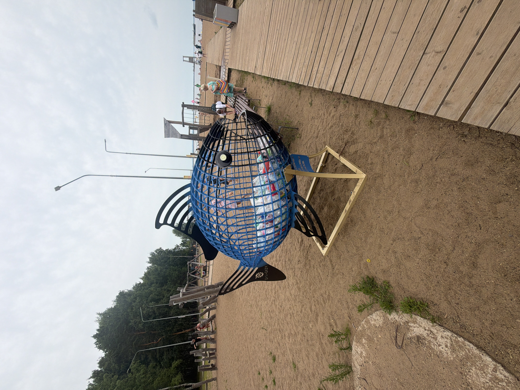 This image captures an outdoor scene on a sandy beach, likely in the city of Chaikovsky, Russia, given the context provided by the prompt. The central focus is a large, blue, fish-shaped sculpture made of wire mesh. Inside the fish's body, numerous plastic bottles are visible, suggesting it may be a recycling bin or art installation promoting waste reduction.
The sculpture is placed on the sand, supported by a light-colored metal frame. To the right of the sculpture, a wooden boardwalk runs parallel to the beach, made of light-colored planks. In the background, a playground with wooden structures and swings is visible, indicating a public recreational area.
Several people are present in the scene. Some are walking along the boardwalk or sitting on a bench near the sculpture. Others are visible in the playground area and further down the beach, where the sea or a large body of water is present. The sky is overcast with a cloudy, pale grey appearance. Trees line the edge of the sandy area behind the playground.
The overall impression is one of a public beach or park area with an artistic and environmental theme.
