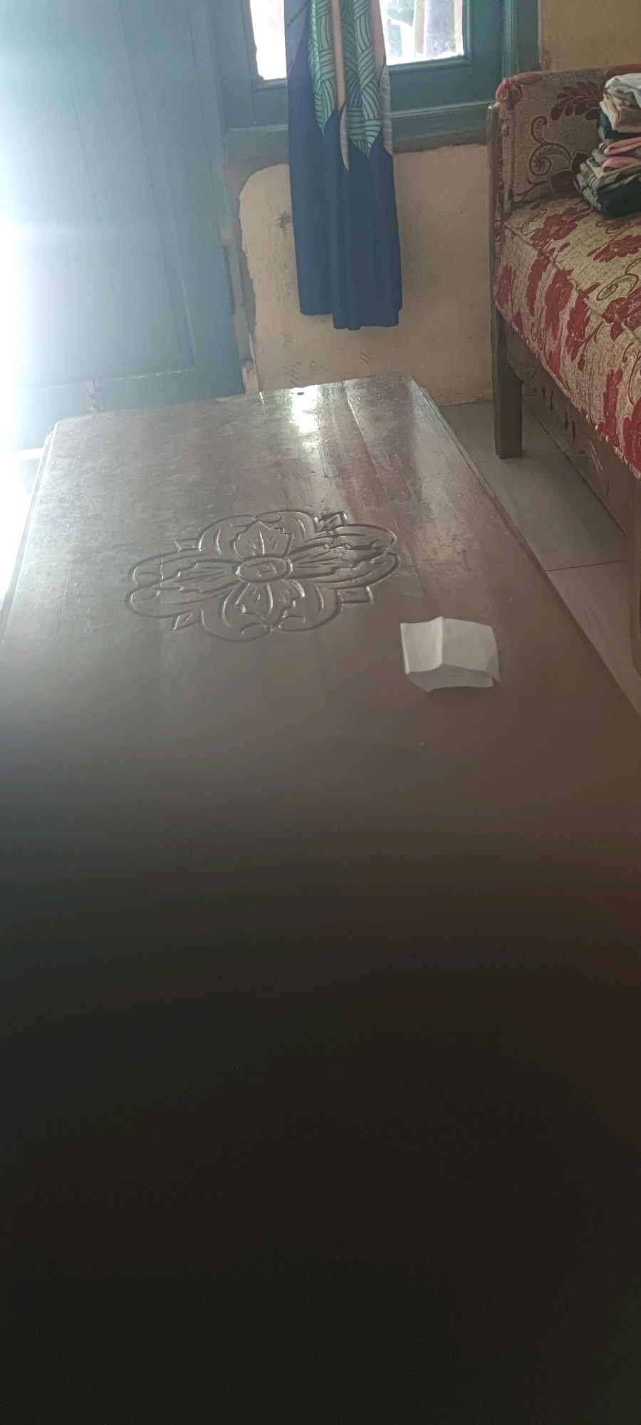 The image shows a close-up of a wooden table with a carved floral design. A small piece of white paper is placed on the table near the carving. In the background, a couch with a red and white patterned fabric is visible, as well as a window with blue and green curtains. The scene appears to be indoors, possibly in a residential setting. There is a strong light source coming from the left, casting a glare on the table. The setting is described as being in Gambarsari, Indonesia.