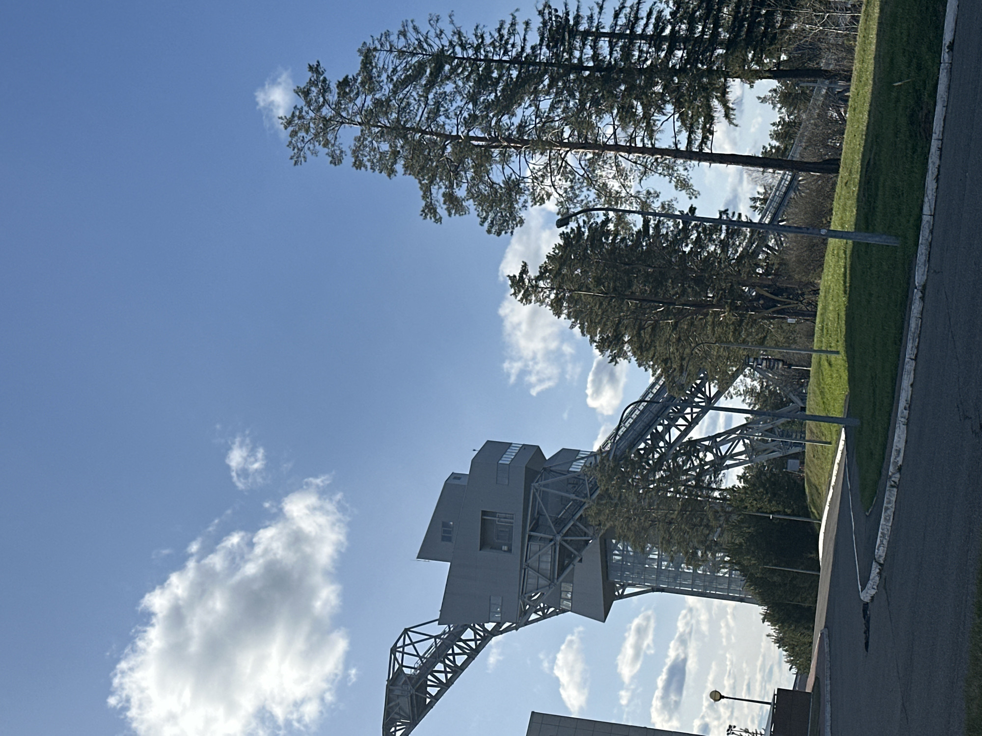 The image shows a sideways view of a port crane located in Прикамский, Russia. The crane is gray with a lattice structure and a box-like cabin at the top. The sky is blue with scattered clouds. Tall pine trees are visible in the background. A paved path runs along the right side of the image, bordering a grassy area.