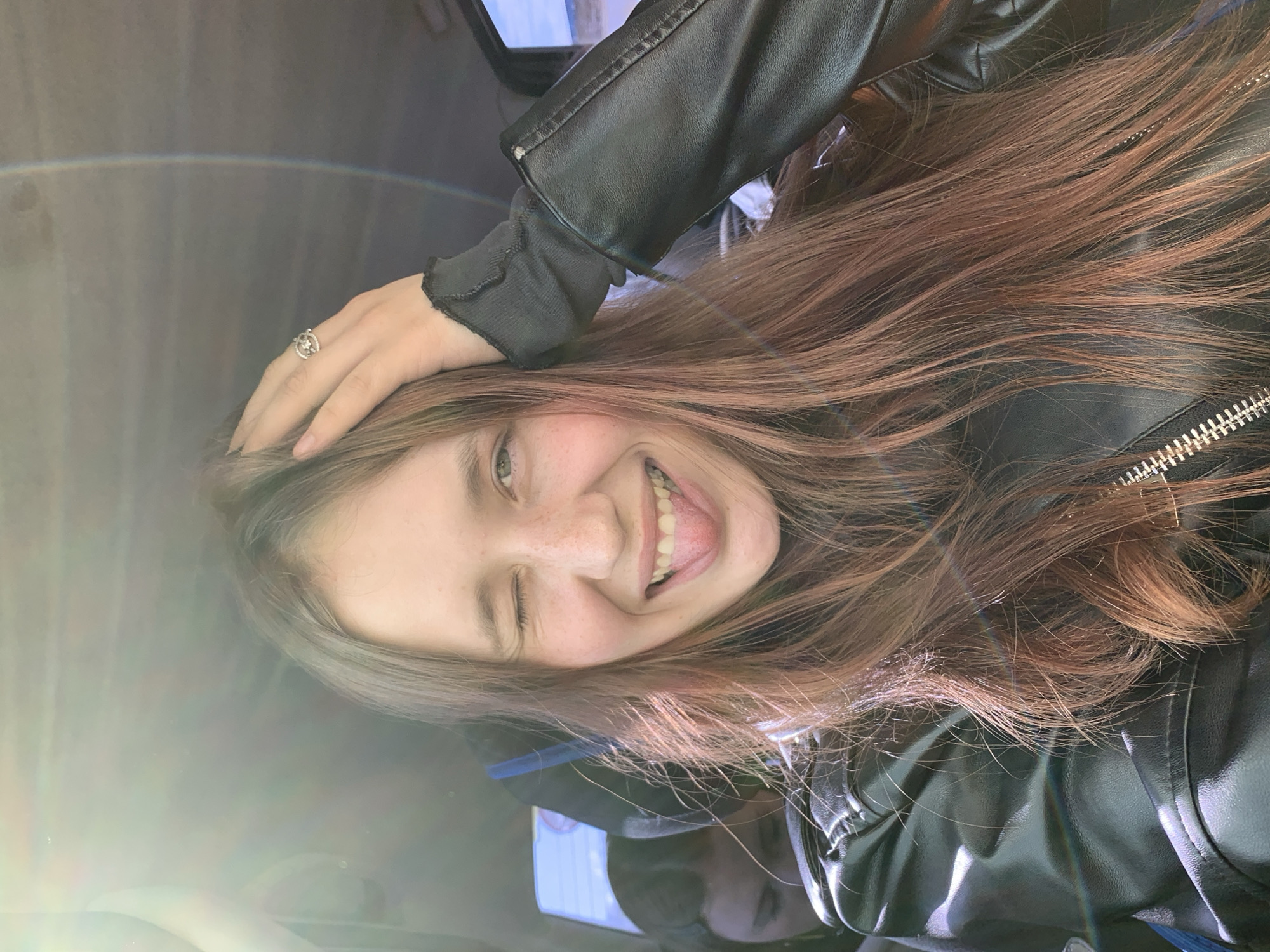 A close-up selfie captures a young person, likely female, with long brown hair, winking with their right eye and smiling broadly with their tongue slightly out, conveying a playful and cheerful mood. They are wearing a black leather jacket over a dark, long-sleeved ribbed top. A ring with a circular design is visible on the ring finger of their right hand, which is resting on their head. Bright, possibly natural light, enters from the left side of the frame, creating a strong lens flare effect and a faint rainbow arc across the upper left quadrant of the image. The background is blurred but appears to show reflections, possibly from a window or a screen, suggesting an indoor setting like a car interior or a room. No discernible text is visible in the image. The provided location context is Гаревая, Russia.